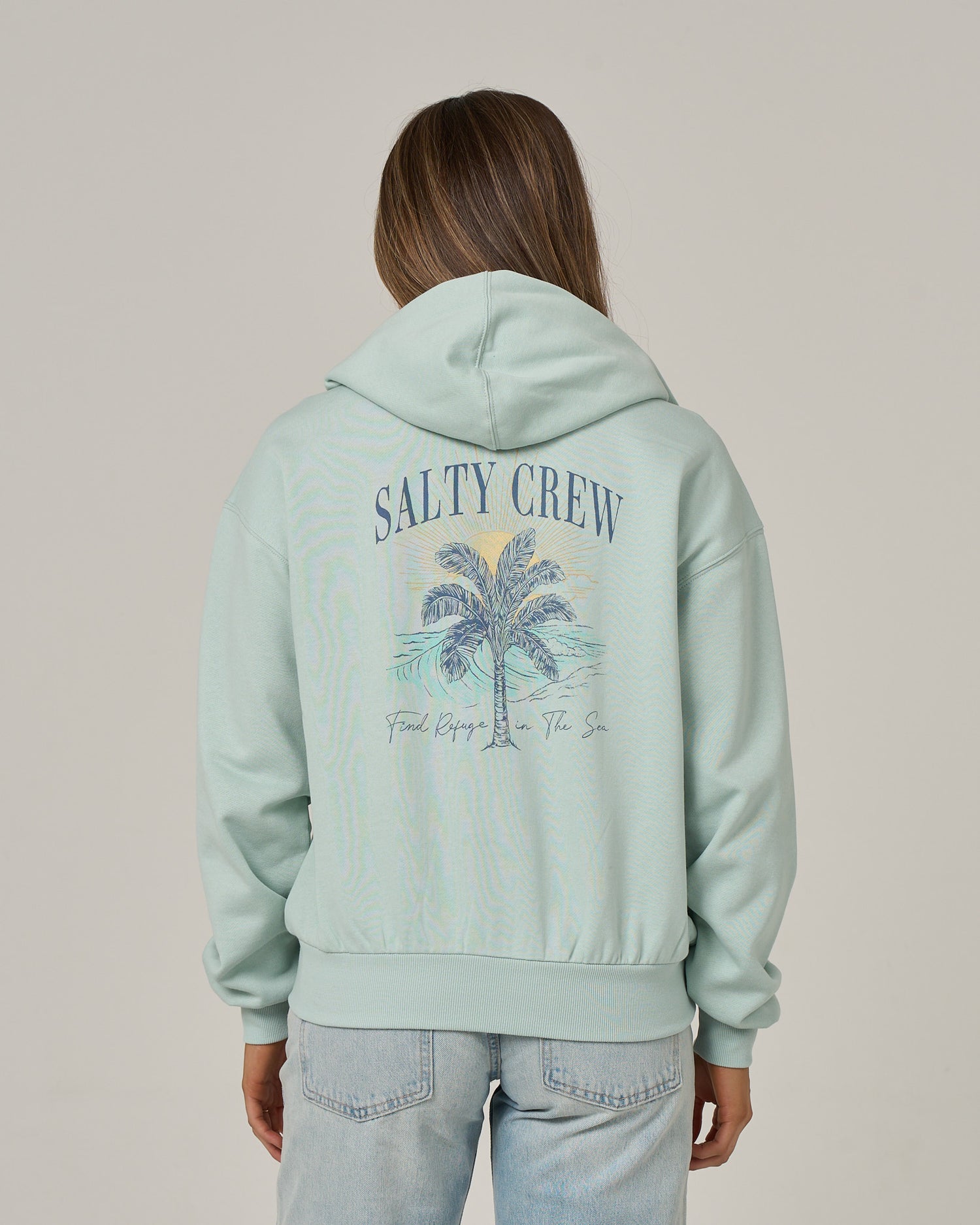 A person is wearing the Salty Crew Pretty Palm Hooded Zip in Cloud Blue, featuring a zip-front design and a palm tree graphic with “Find Refuge In The Sea” text on the back. This hoodie is crafted from comfortable cotton-polyester fleece and is showcased against a plain background.