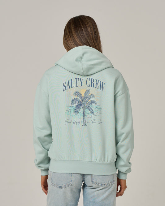 A person is wearing the Salty Crew Pretty Palm Hooded Zip in Cloud Blue, featuring a zip-front design and a palm tree graphic with “Find Refuge In The Sea” text on the back. This hoodie is crafted from comfortable cotton-polyester fleece and is showcased against a plain background.