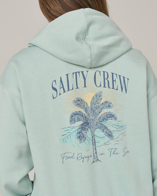 A person wears the Salty Crew Pretty Palm Hooded Zip in Cloud Blue, featuring a relaxed fit and a back graphic of a palm tree, ocean waves, and the sun alongside “SALTY CREW” and “Find Refuge in The Sea” printed on it. Crafted from soft cotton-polyester fleece for comfort and warmth.