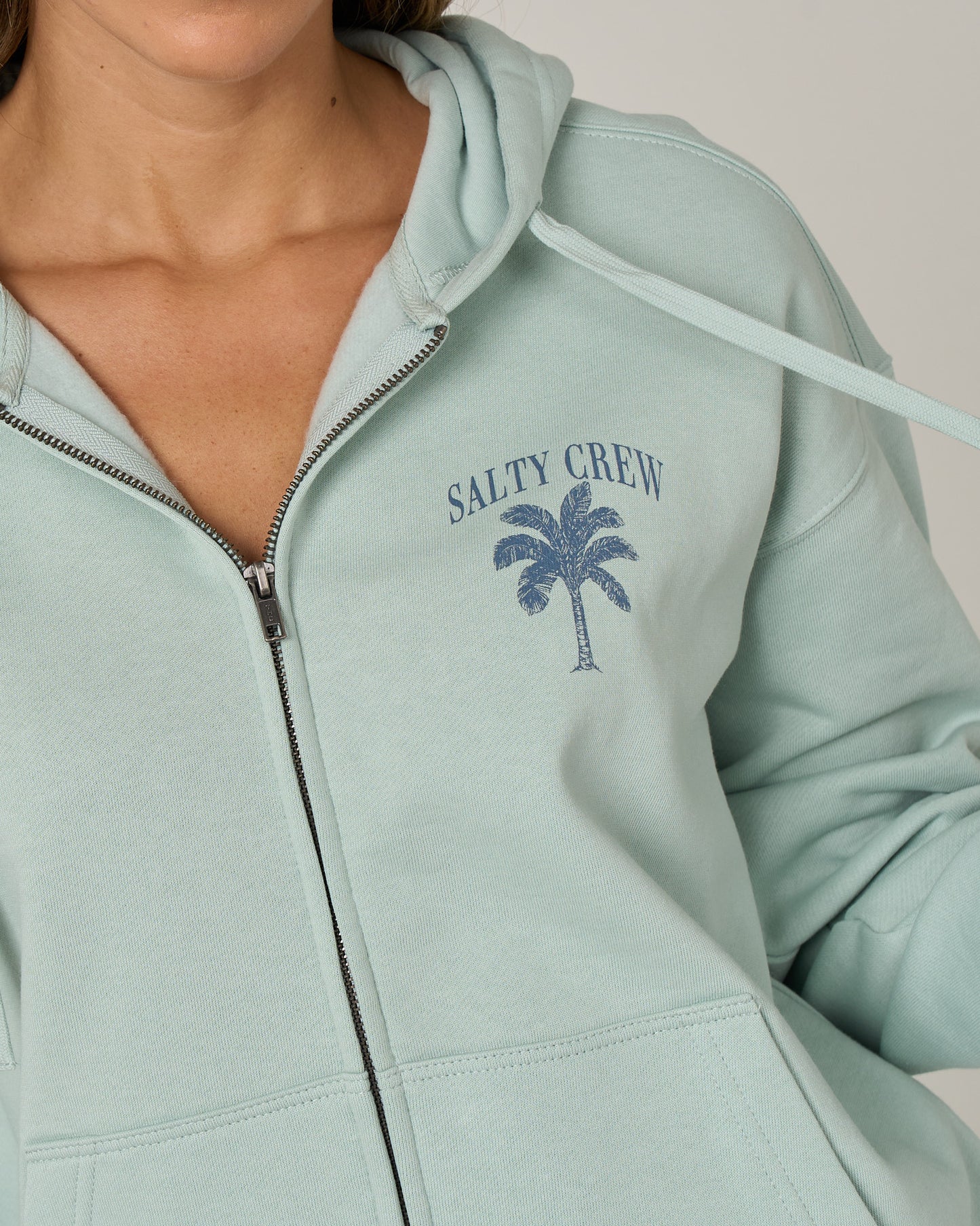 A person is wearing the Salty Crew Pretty Palm Hooded Zip in Cloud Blue—a relaxed fit, zip-front hoody featuring a palm tree graphic and Salty Crew branding on the chest. Made from soft cotton polyester fleece, only the lower half of their face and torso are visible.