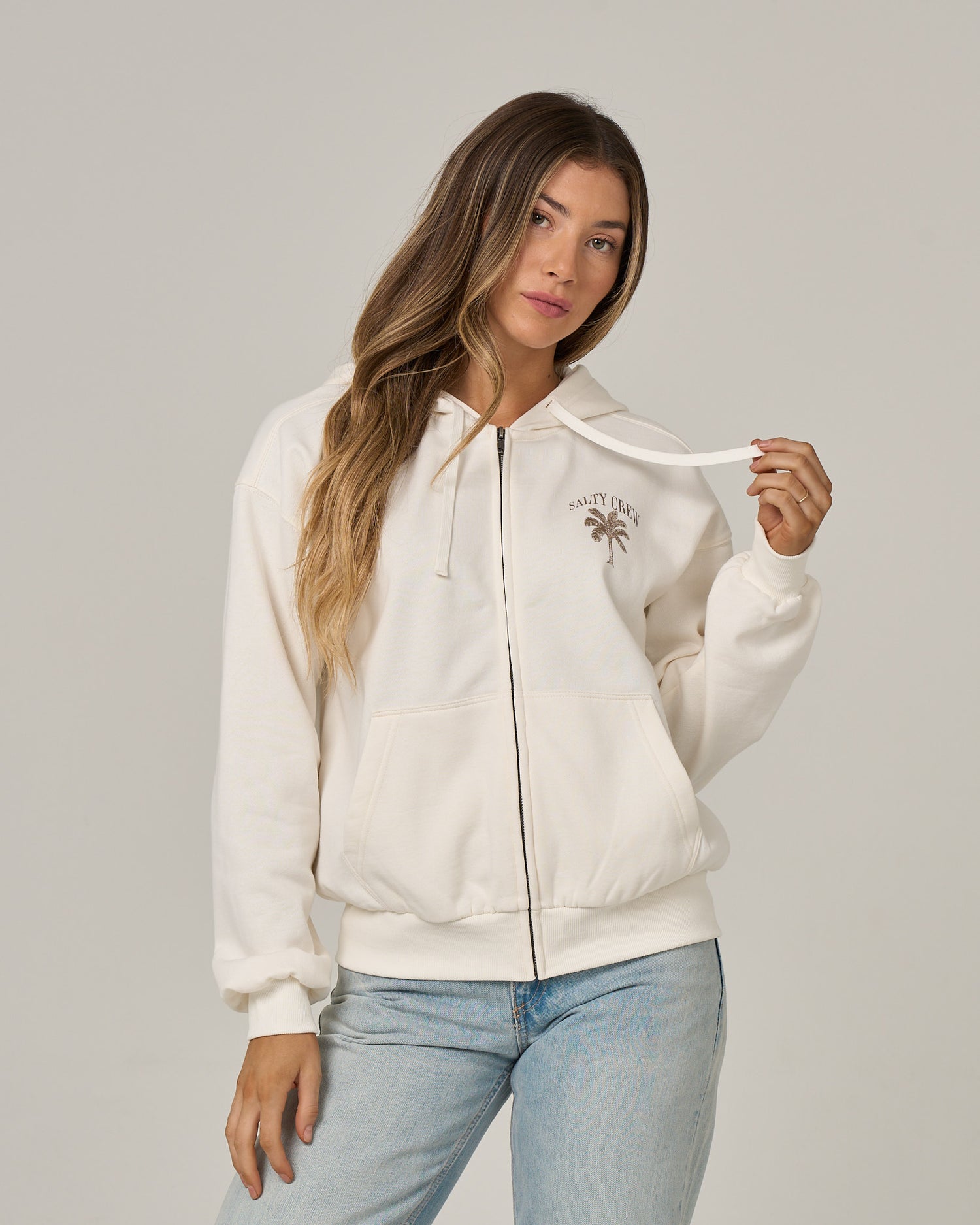 A woman with long, wavy hair stands against a plain background, holding one drawstring of the Salty Crew Pretty Palm Hooded Zip in Off White. This relaxed fit hoody features a small palm tree graphic and Salty Crew text on the chest, a classic kangaroo pocket, and is crafted from a soft cotton-polyester fleece.