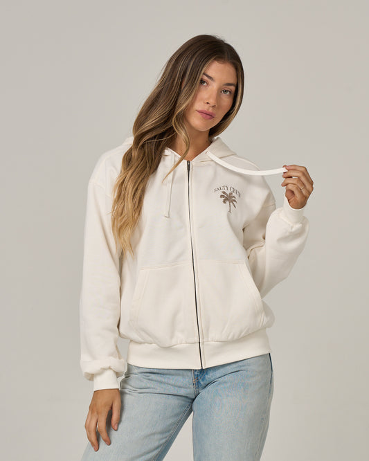 A woman with long, wavy hair stands against a plain background, holding one drawstring of the Salty Crew Pretty Palm Hooded Zip in Off White. This relaxed fit hoody features a small palm tree graphic and Salty Crew text on the chest, a classic kangaroo pocket, and is crafted from a soft cotton-polyester fleece.