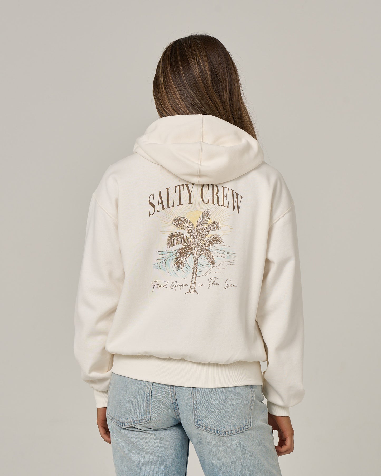 A person wearing the Salty Crew Pretty Palm Hooded Zip in Off White—a relaxed fit zip hoody featuring a palm tree graphic and the words “SALTY CREW” and “Find Refuge in the Sea” on the back, crafted from soft cotton-polyester fleece with a convenient kangaroo pocket—is paired with light blue jeans and stands facing away.