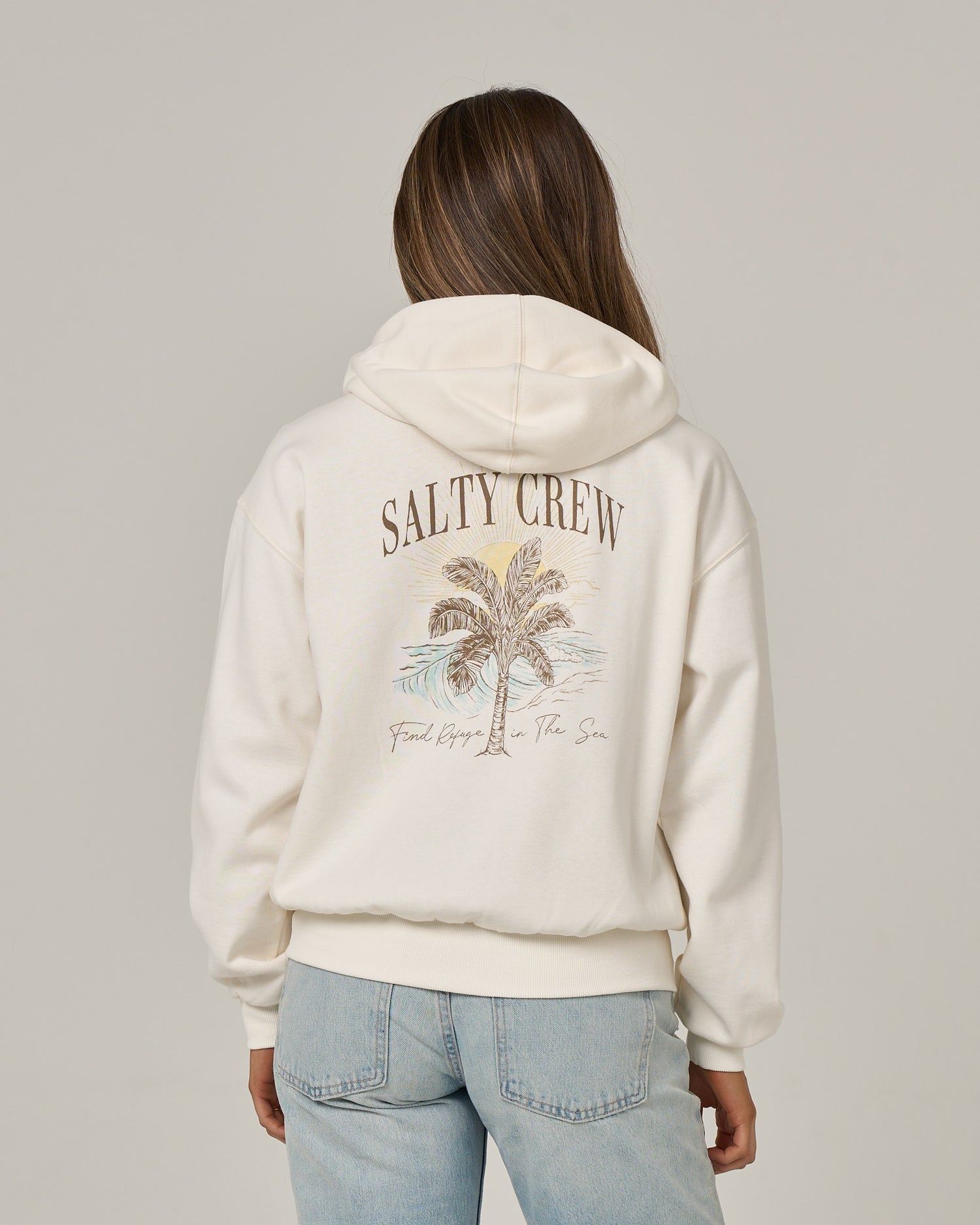 A person wearing the Salty Crew Pretty Palm Hooded Zip in Off White—a relaxed fit zip hoody featuring a palm tree graphic and the words “SALTY CREW” and “Find Refuge in the Sea” on the back, crafted from soft cotton-polyester fleece with a convenient kangaroo pocket—is paired with light blue jeans and stands facing away.