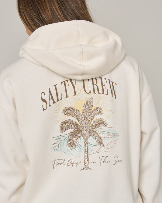 A person wearing the Salty Crew Pretty Palm Hooded Zip in Off White—a relaxed fit cotton-polyester fleece zip hoody featuring “SALTY CREW” above a palm tree and ocean wave graphic, with “Find Refuge in The Sea” written underneath on the back, finished with a cozy kangaroo pocket.