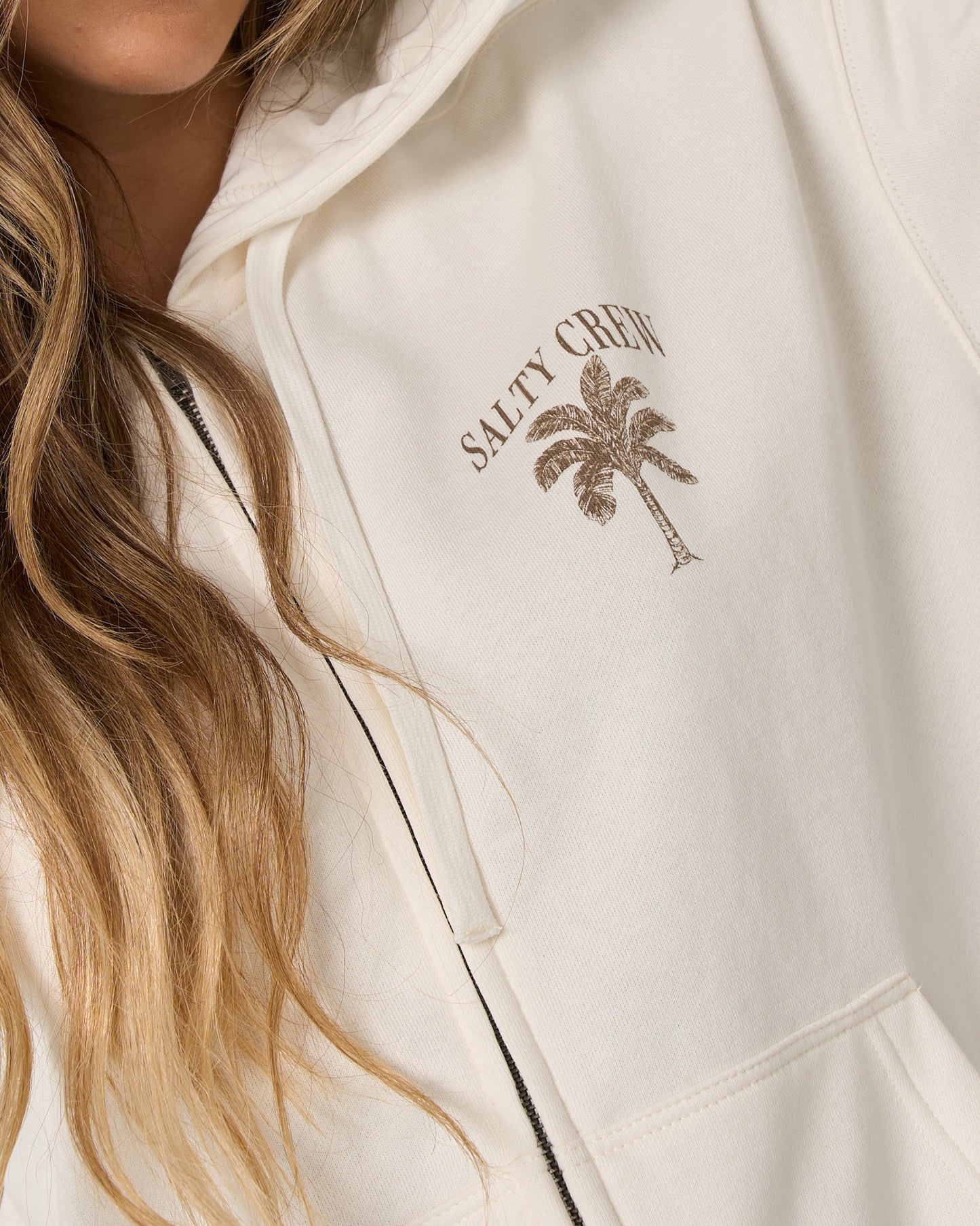 A person wearing the Salty Crew Pretty Palm Hooded Zip in off white, a relaxed fit hoodie featuring a palm tree and SALTY CREW graphic on the chest. Crafted from soft cotton-polyester fleece, this white zip-up includes a classic kangaroo pocket. Only the lower part of their face and their long wavy hair are visible.