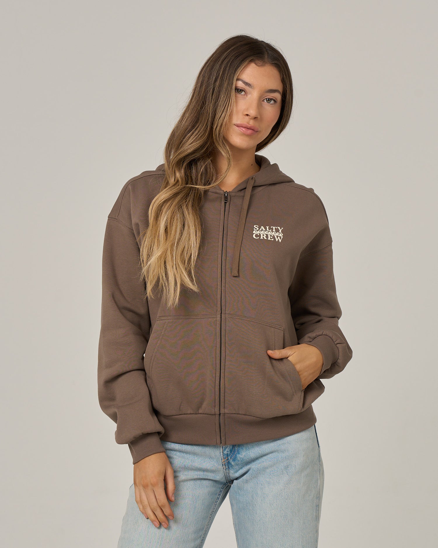 A model with long brown hair poses against a light gray background wearing the Salty Crew Lobster Roll Hooded Zip in Washed Brown—a soft cotton polyester fleece zip-up featuring white SALTY CREW chest print—styled with light-wash blue jeans for a relaxed fit.