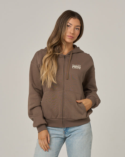A model with long brown hair poses against a light gray background wearing the Salty Crew Lobster Roll Hooded Zip in Washed Brown—a soft cotton polyester fleece zip-up featuring white SALTY CREW chest print—styled with light-wash blue jeans for a relaxed fit.