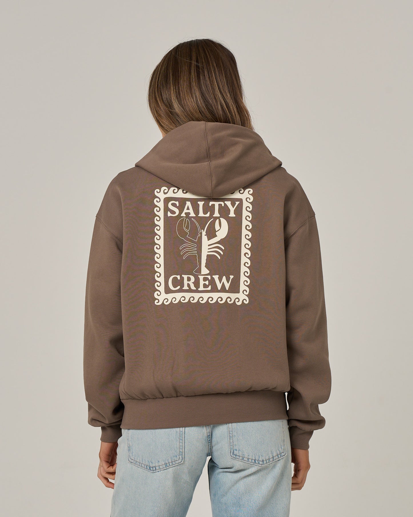 A person wearing the Salty Crew Lobster Roll Hooded Zip in washed brown—a relaxed fit zip-front hoody featuring a lobster graphic on the back, crafted from soft cotton-polyester fleece—paired with light blue jeans and standing against a plain background.