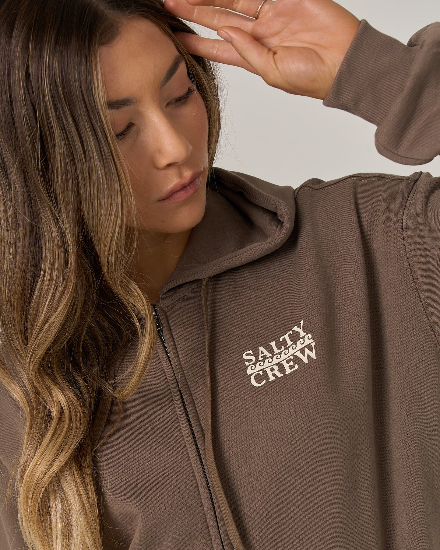 Wearing the Salty Crew Lobster Roll Hooded Zip in Washed Brown, a person with long, wavy hair raises their hand near their head and looks down. Crafted from a comfortable cotton-polyester fleece and featuring Salty Crew branding on the chest, this relaxed fit hoodie offers both style and ease.