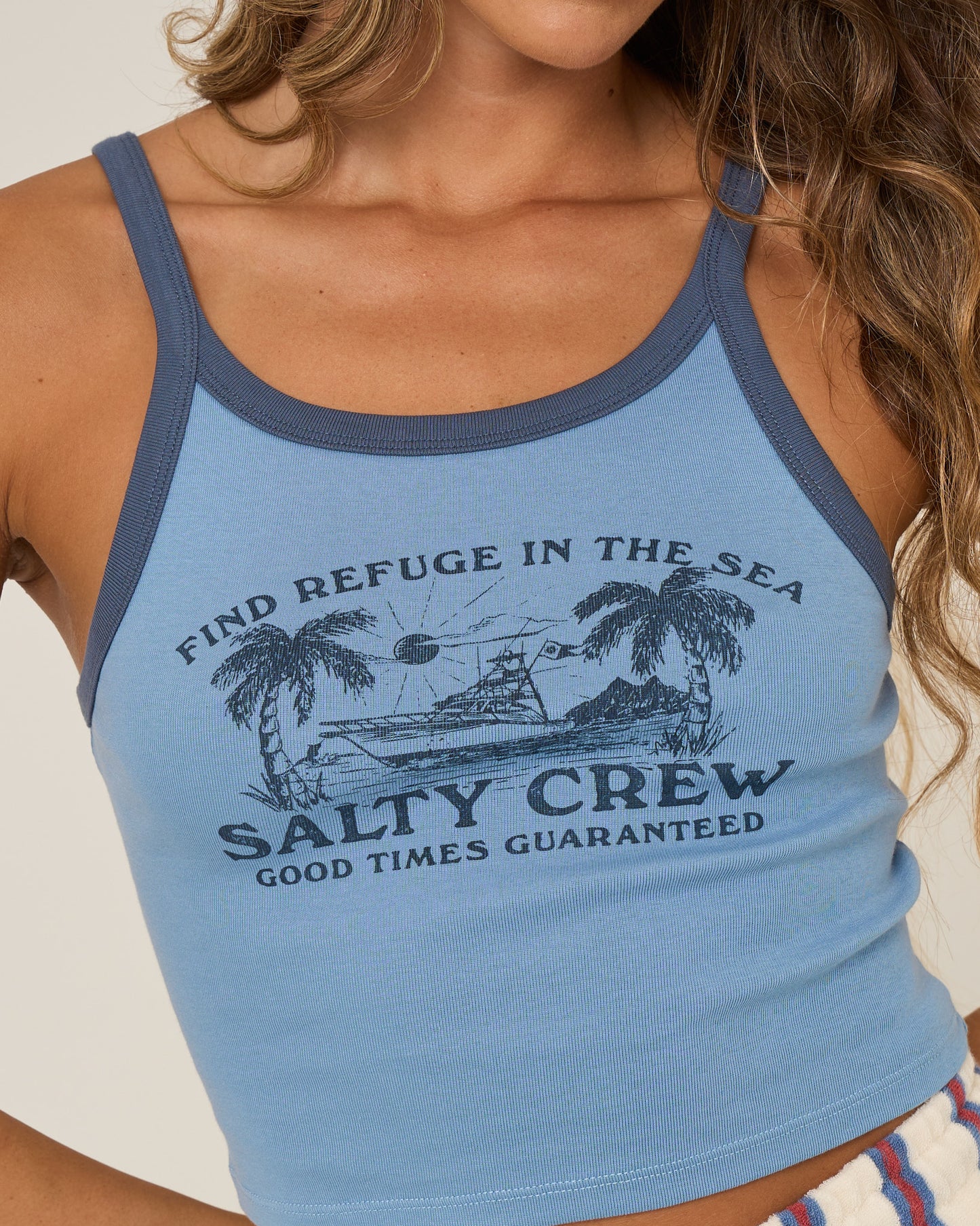 A woman wears the Salty Crew Charter Tank in Ash Blue, a 100% cotton rib tank with navy trim, featuring palm trees, a ship graphic, and the text “Find Refuge in the Sea. Salty Crew. Good Times Guaranteed.”.