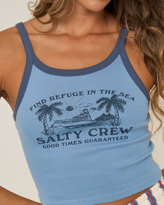A woman wears the Salty Crew Charter Tank in Ash Blue, a 100% cotton rib tank with navy trim, featuring palm trees, a ship graphic, and the text “Find Refuge in the Sea. Salty Crew. Good Times Guaranteed.”.