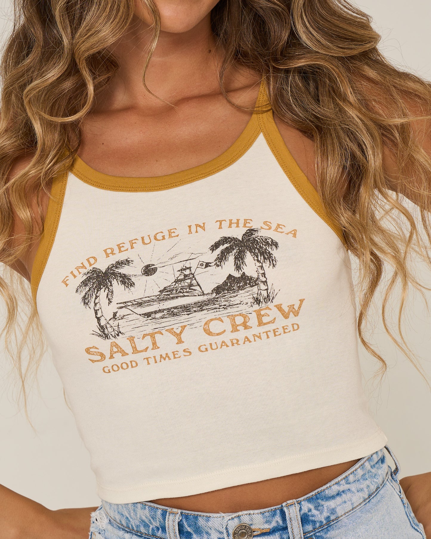 A woman with wavy hair wears the Salty Crew Charter Tank in off white—a sleeveless, high neck top featuring mustard trim, a beach-themed print, and "Find refuge in the sea, Salty Crew, Good times guaranteed"—paired with light blue jeans.