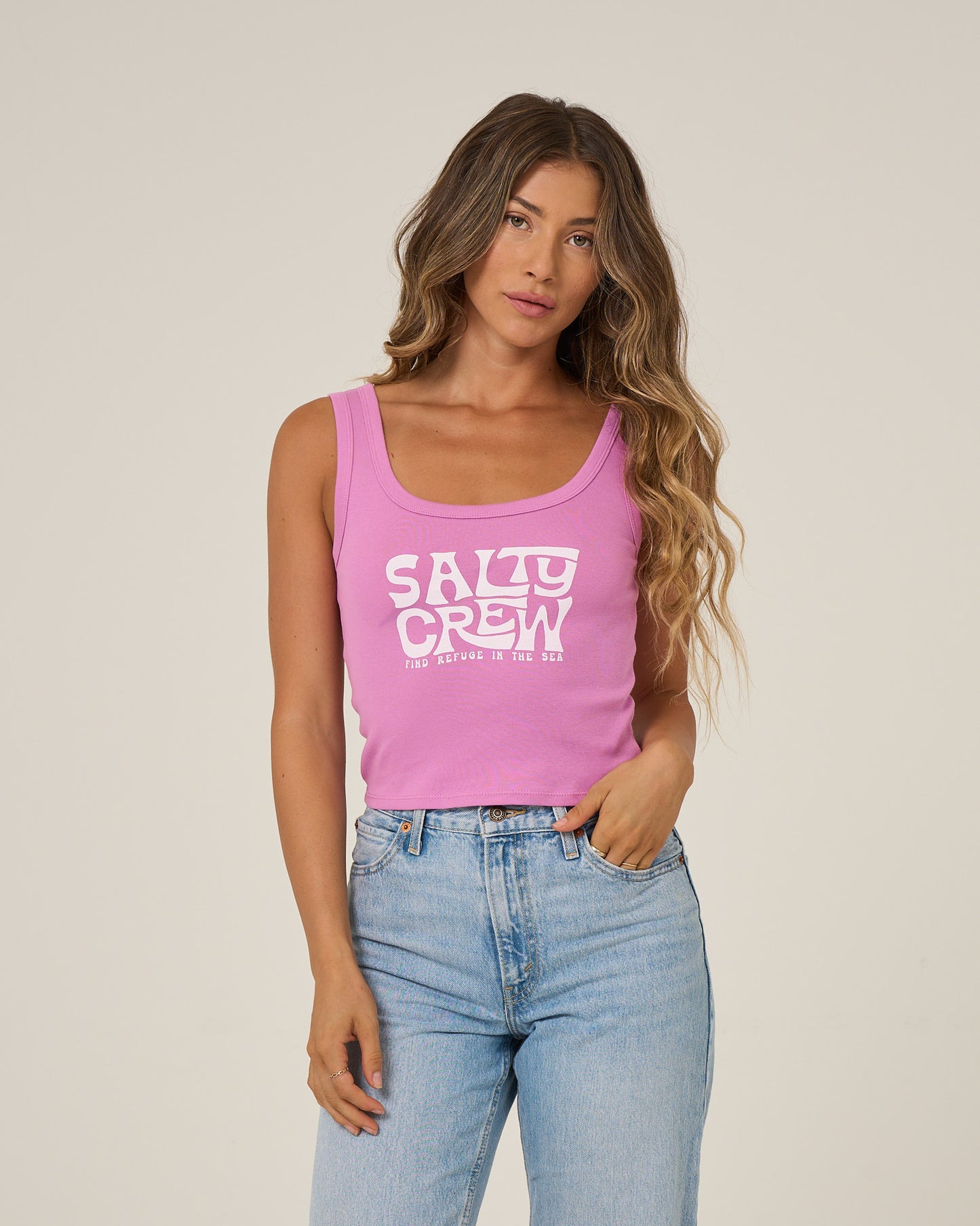 A woman with long wavy hair stands against a beige background wearing the Salty Crew Freestyle Tank in violet and light blue jeans, one hand in her pocket.