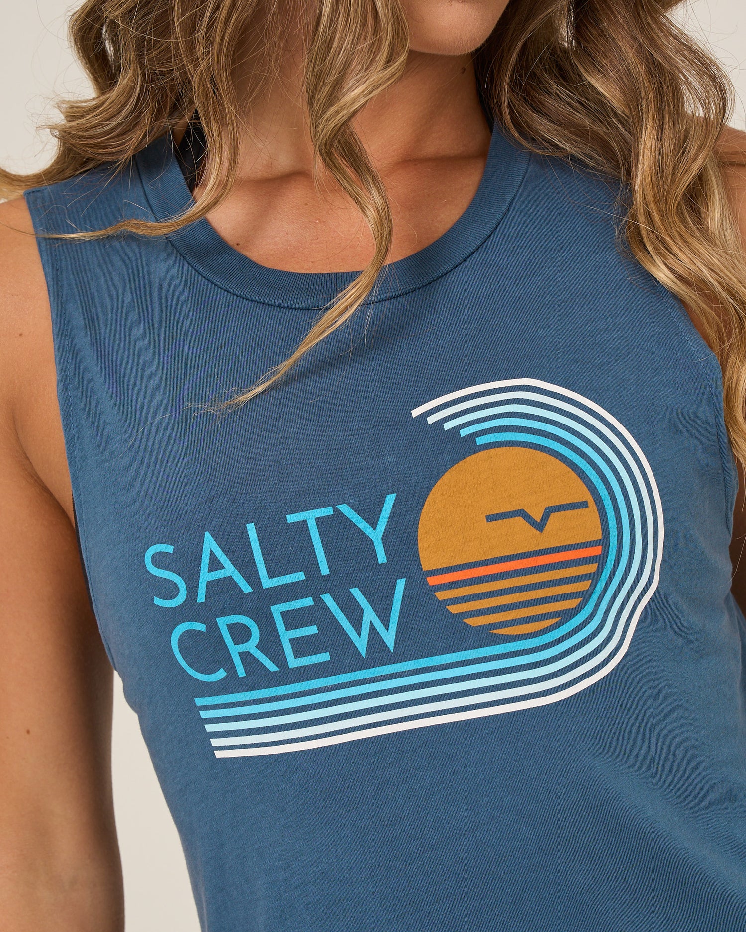 A person wearing the Salty Crew Ombre Wave Muscle Tank in Midnight Navy, featuring stylized wave and orange sun graphics, Salty Crew text on the front, and side slits for comfort.