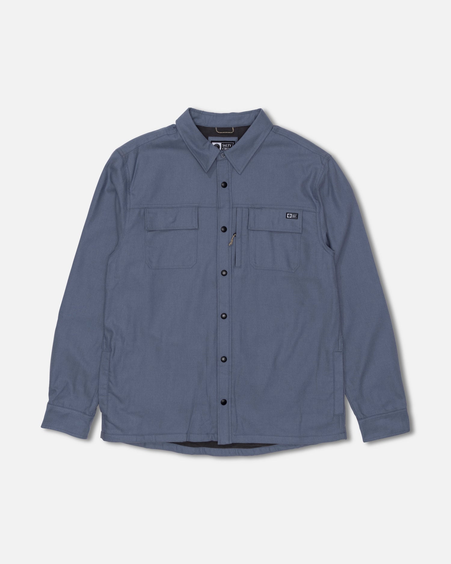 The Salty Crew Fathom Tech Shacket in Slate Grey is a long-sleeve, button-up shirt crafted from mid-weight twill for versatile wear. It features a classic collar, black buttons, and two chest pockets—one with a zipper detail—and is displayed on a plain white background.