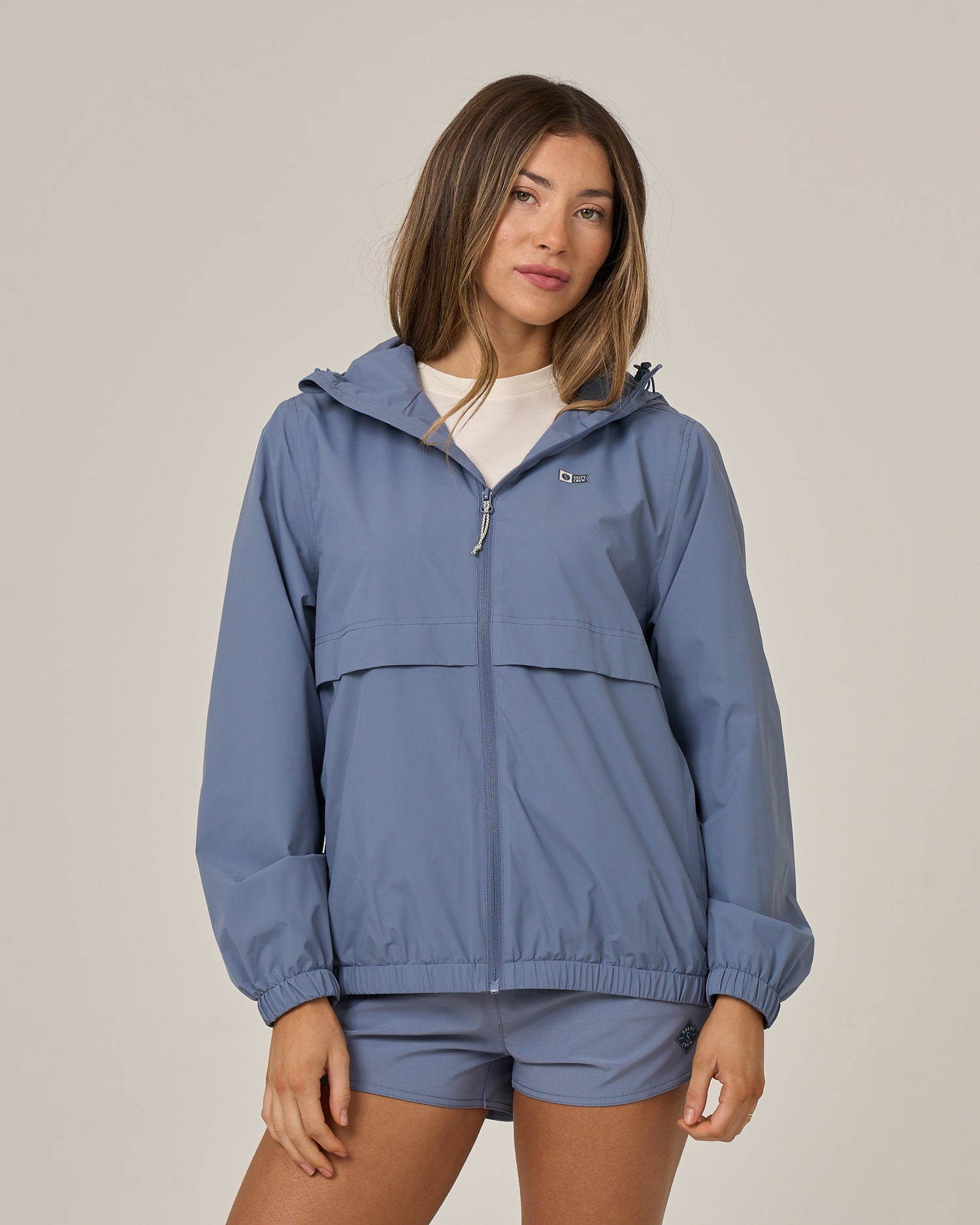 A woman wearing the Salty Crew Barrier Windbreaker in Flint, featuring a cinching system, and matching shorts stands against a plain background, looking toward the camera with a neutral expression.