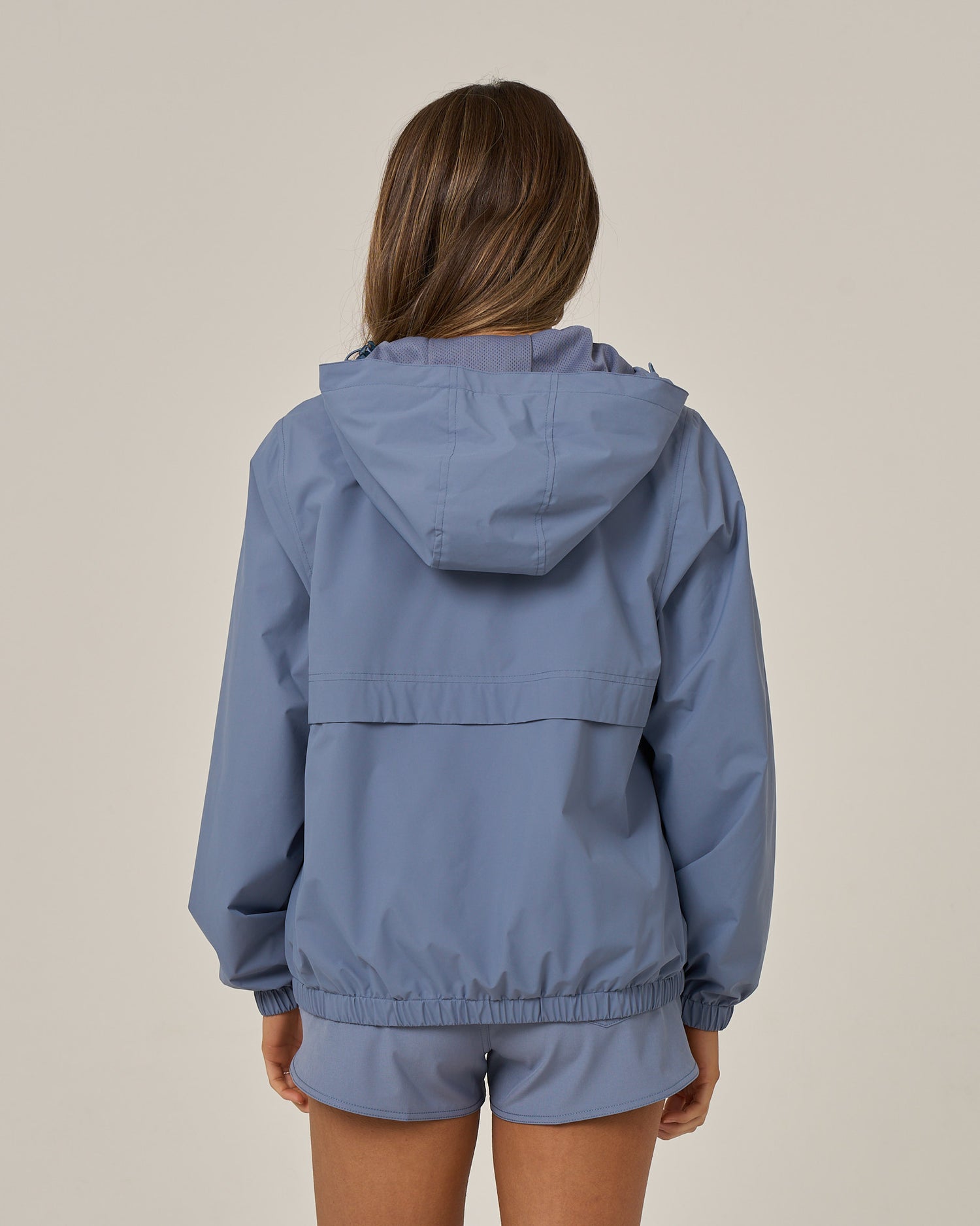 A person with long brown hair is seen from the back, standing against a plain light background, wearing the Salty Crew Barrier Windbreaker in Flint—a light blue, water-resistant jacket featuring a hood with cinching system—and matching shorts.