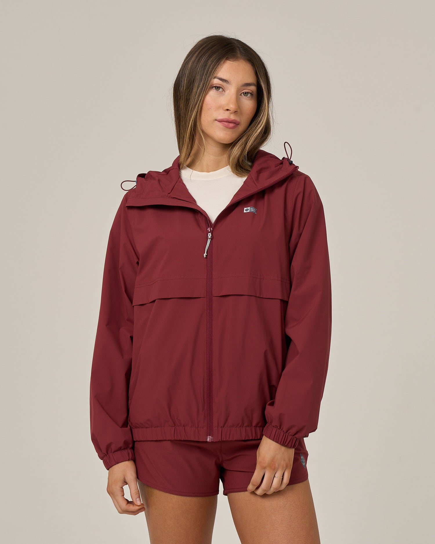 Wearing the Salty Crew Barrier Windbreaker in maroon and matching shorts, a woman stands against a plain light background, looking at the camera with a neutral expression.