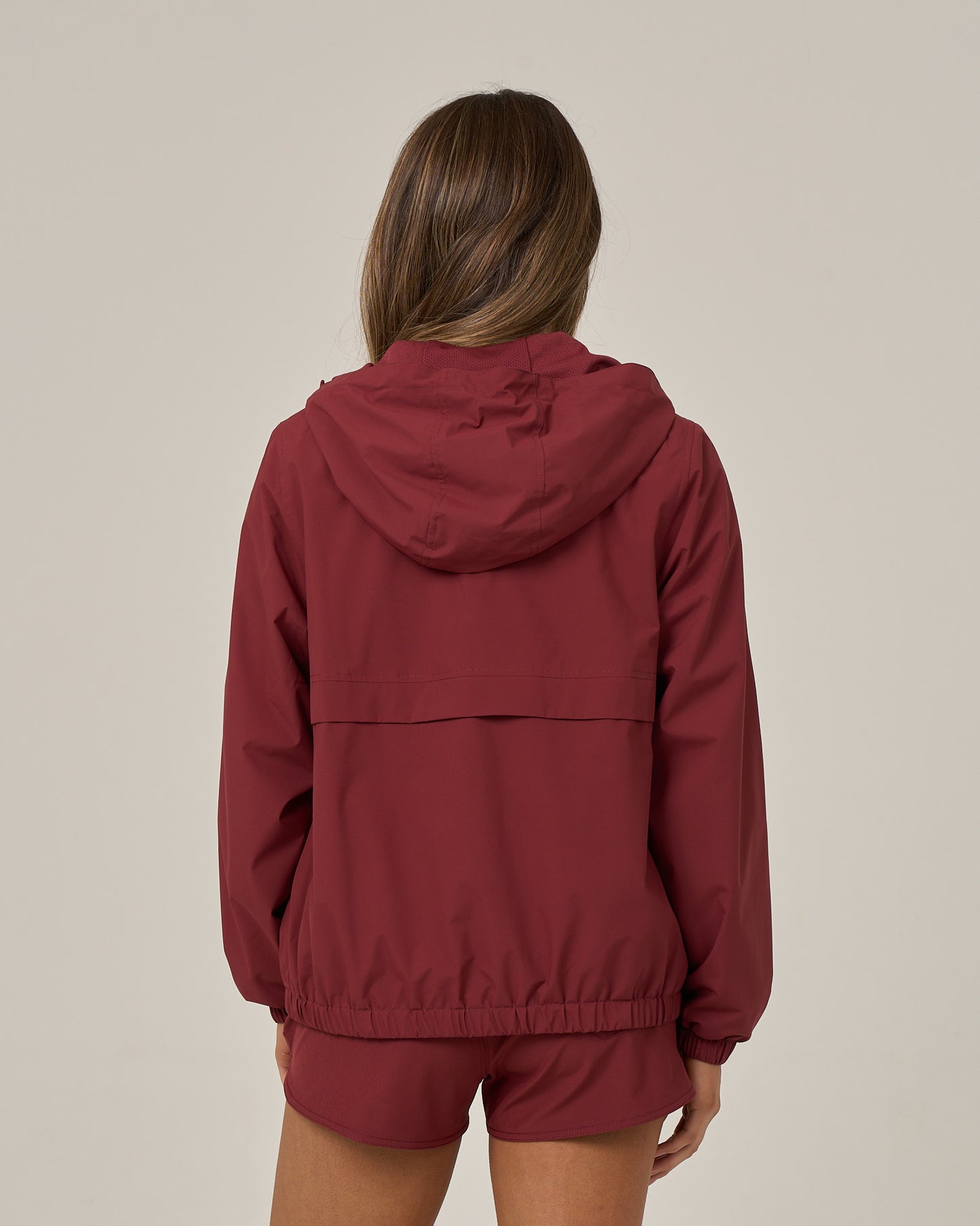 A person with long brown hair, seen from behind, wears the Salty Crew Barrier Windbreaker in maroon—crafted from breathable fabric—paired with matching shorts against a plain, light background.