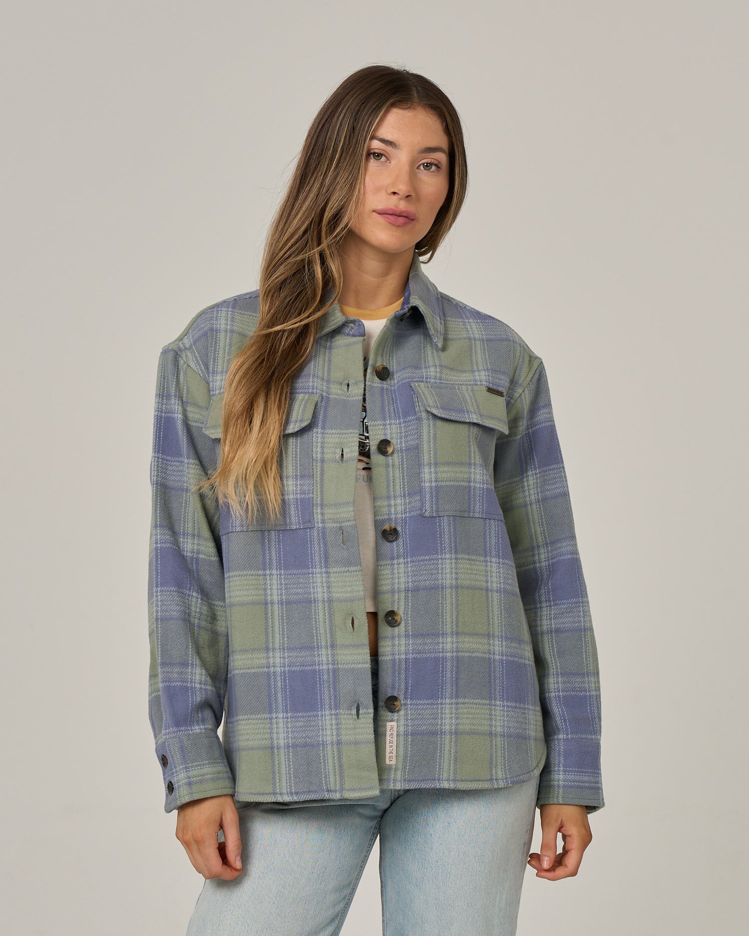 A woman with long brown hair wears the Salty Crew Day Trip Shacket - Flint, crafted from heavyweight cotton blanket flannel in a green and blue plaid pattern, layered over a gray top and light blue jeans as she stands against a plain light background.
