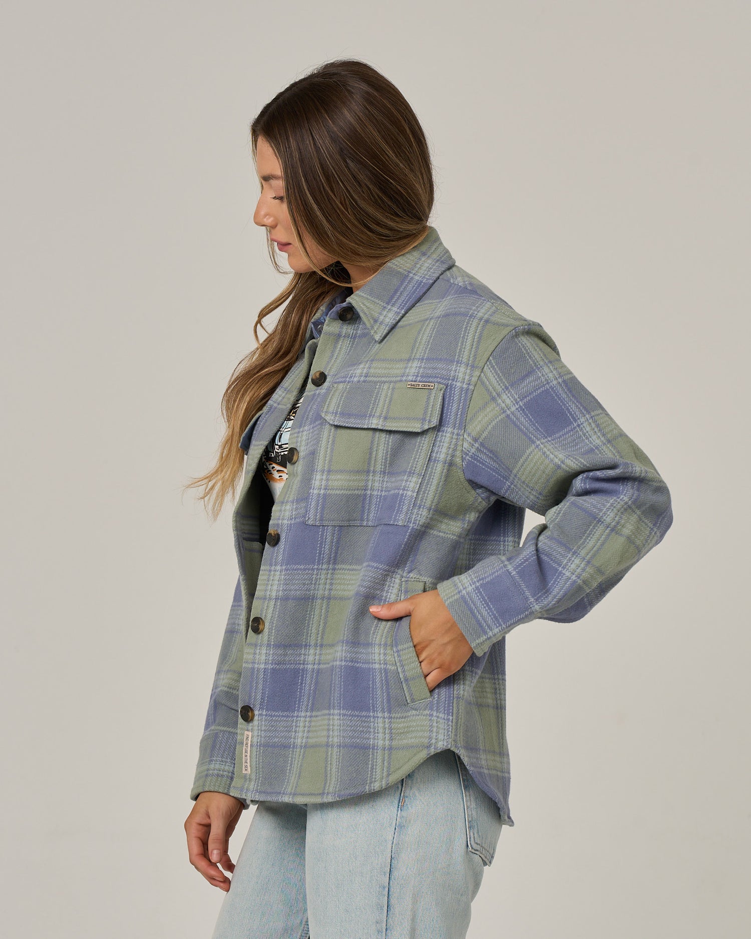 A woman with long brown hair stands sideways against a plain, light background, wearing the Salty Crew Day Trip Shacket in Flint—a heavy-weight, blue and green cotton blanket flannel plaid shirt-jacket—with her left hand in her pocket and paired with light blue jeans.