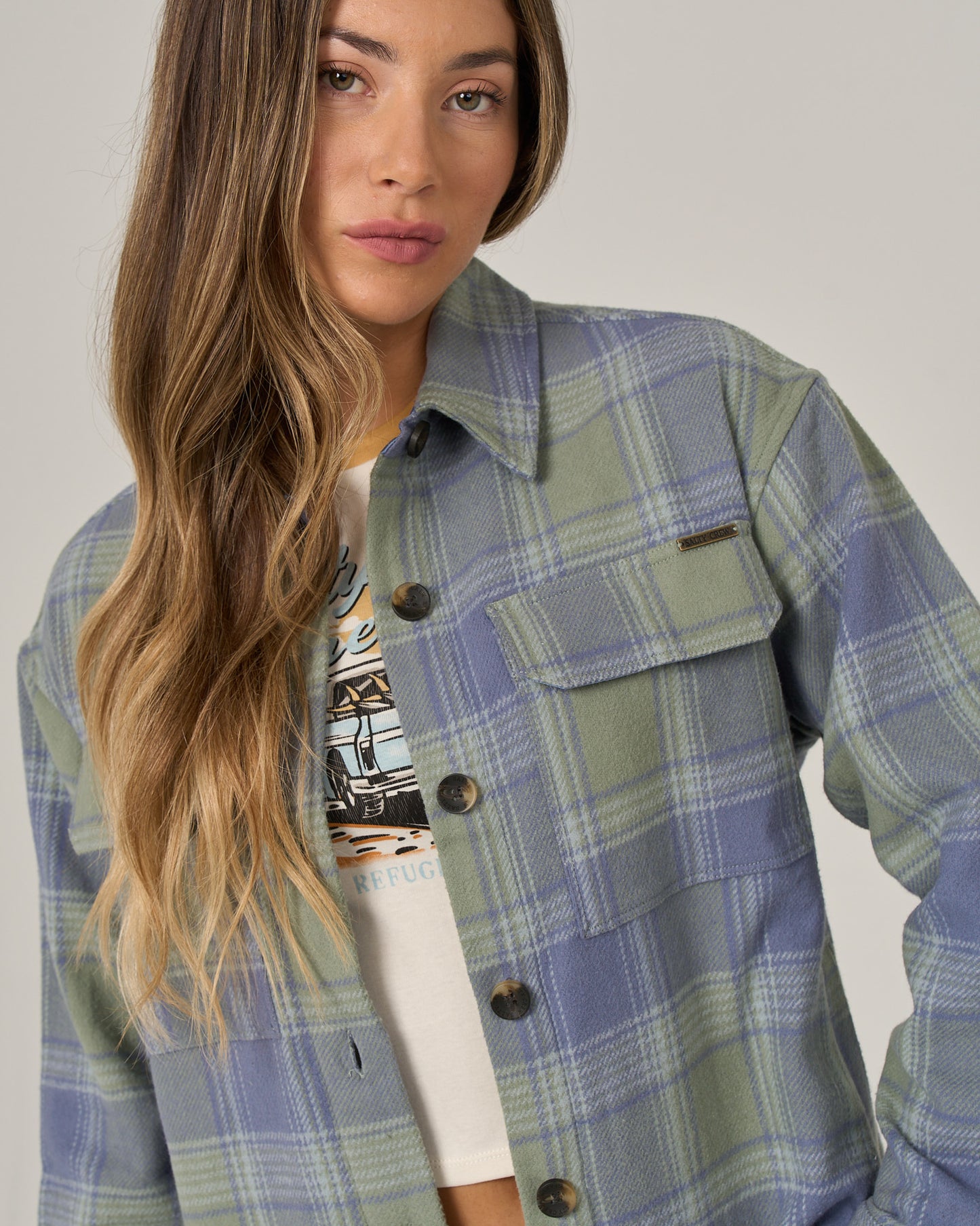 A woman with long, wavy blonde and brown hair stands against a light background, wearing the Salty Crew Day Trip Shacket - Flint in a blue and green plaid over a white printed t-shirt. The heavyweight cotton blanket flannel of the shacket adds warmth and effortless style to her look.