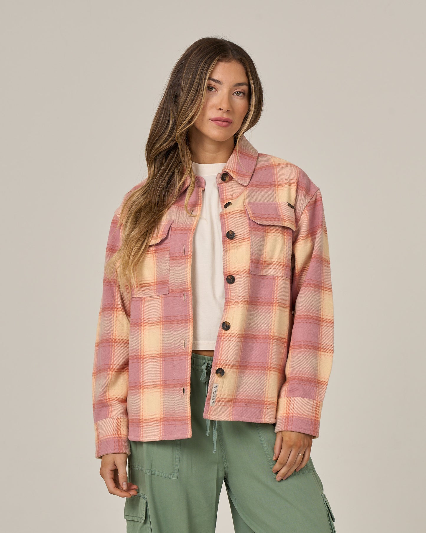 A woman wearing the Salty Crew Day Trip Shacket - Rose, a heavyweight plaid cotton flannel jacket with button closure, over a white shirt and green cargo pants stands against a plain light background, looking at the camera with a neutral expression.