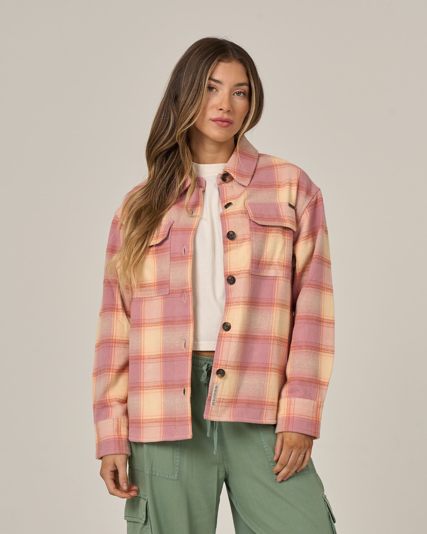 A woman wearing the Salty Crew Day Trip Shacket - Rose, a heavyweight plaid cotton flannel jacket with button closure, over a white shirt and green cargo pants stands against a plain light background, looking at the camera with a neutral expression.
