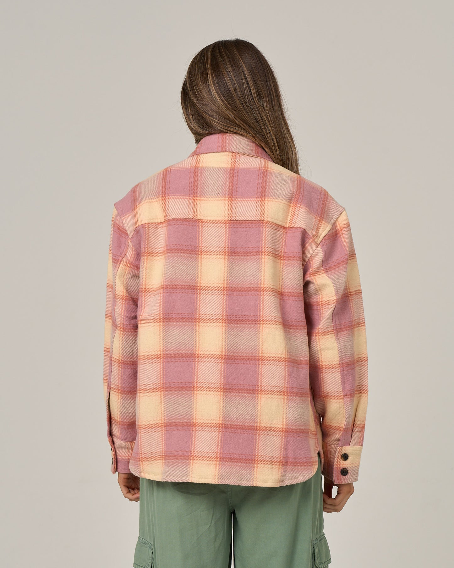A person with long brown hair stands facing away, wearing the Salty Crew Day Trip Shacket - Rose, a heavyweight plaid cotton flannel shacket with button closure in pink and yellow shades, paired with green cargo pants against a plain light background.