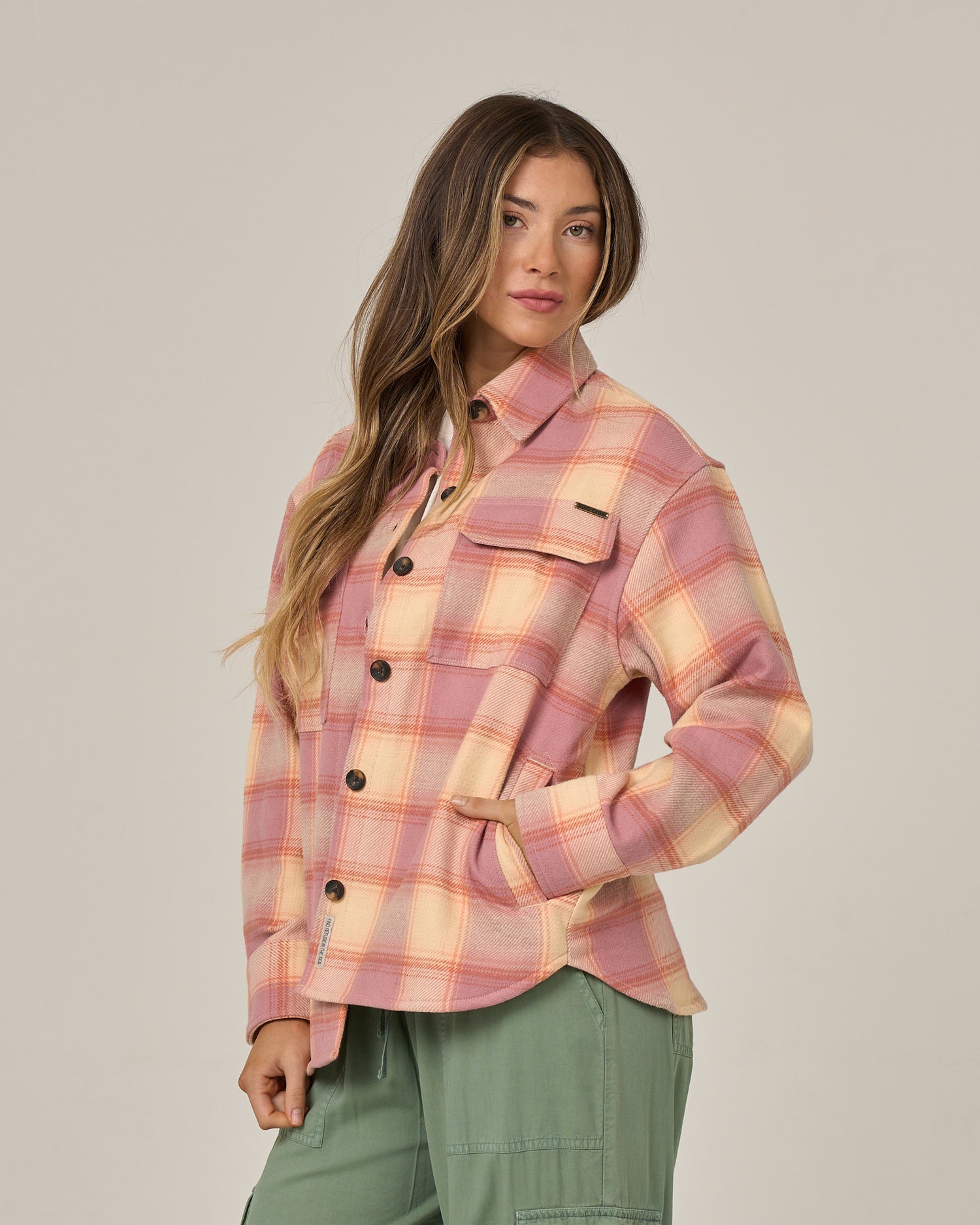 A woman wearing the Salty Crew Day Trip Shacket in Rose—a pink and peach heavyweight plaid cotton flannel with front pockets and button closure—stands with one hand in the pocket of her green pants against a plain background.