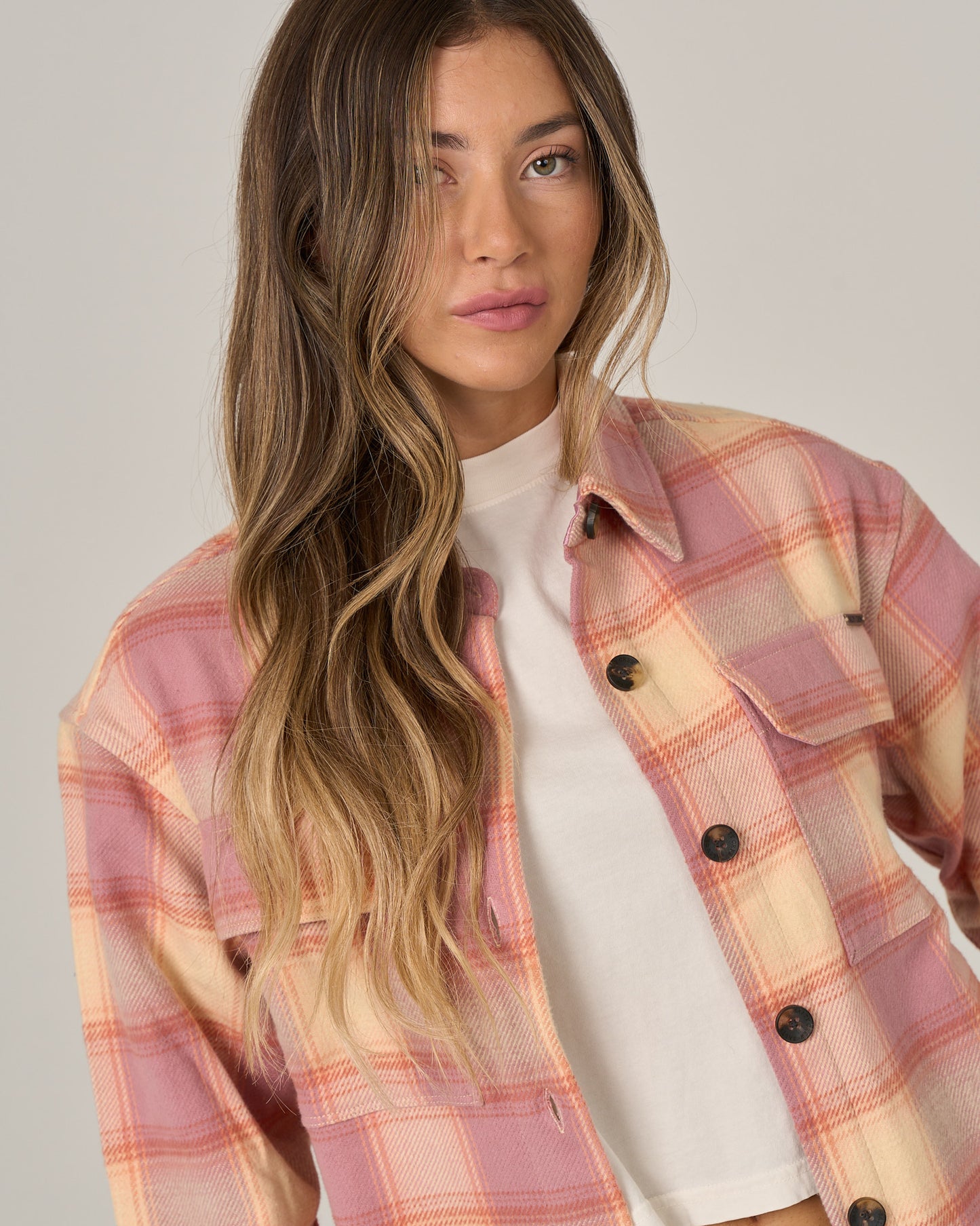 A woman with long, wavy brown hair wears the Salty Crew Day Trip Shacket in Rose—a heavyweight plaid cotton flannel shacket with a button closure—over a white shirt, standing against a plain light background.