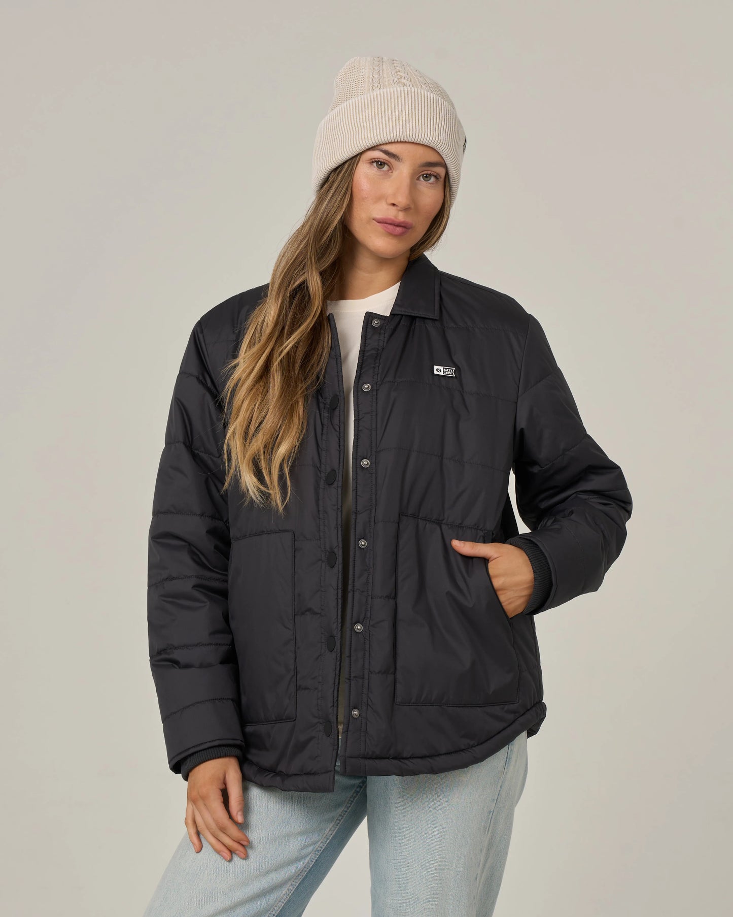 A woman wearing the Salty Crew Barrier Snap Jacket in black, styled with a light knit beanie, white shirt, and light blue jeans, poses with one hand in her jacket pocket against a plain background.