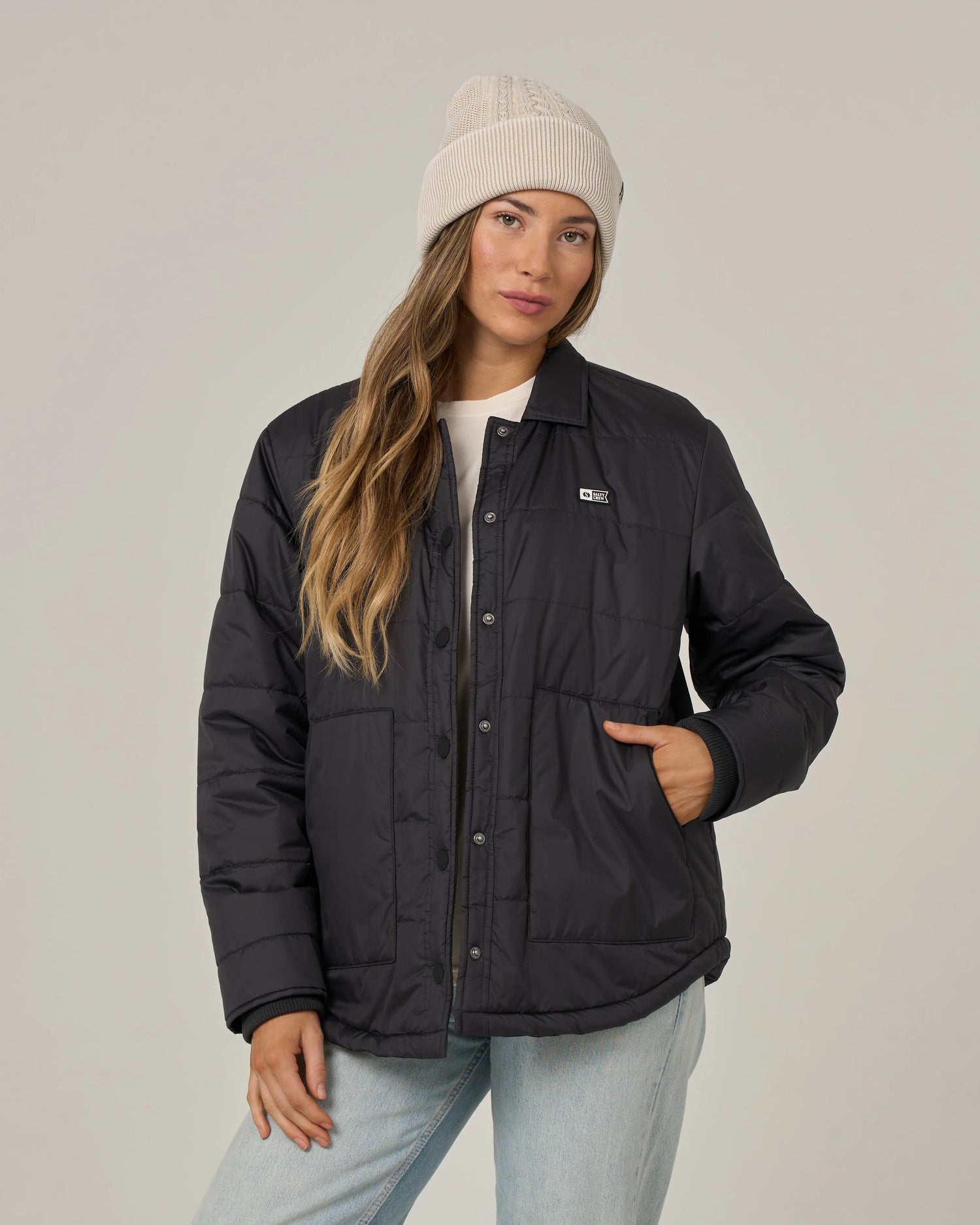 A woman wearing the Salty Crew Barrier Snap Jacket in black, styled with a light knit beanie, white shirt, and light blue jeans, poses with one hand in her jacket pocket against a plain background.