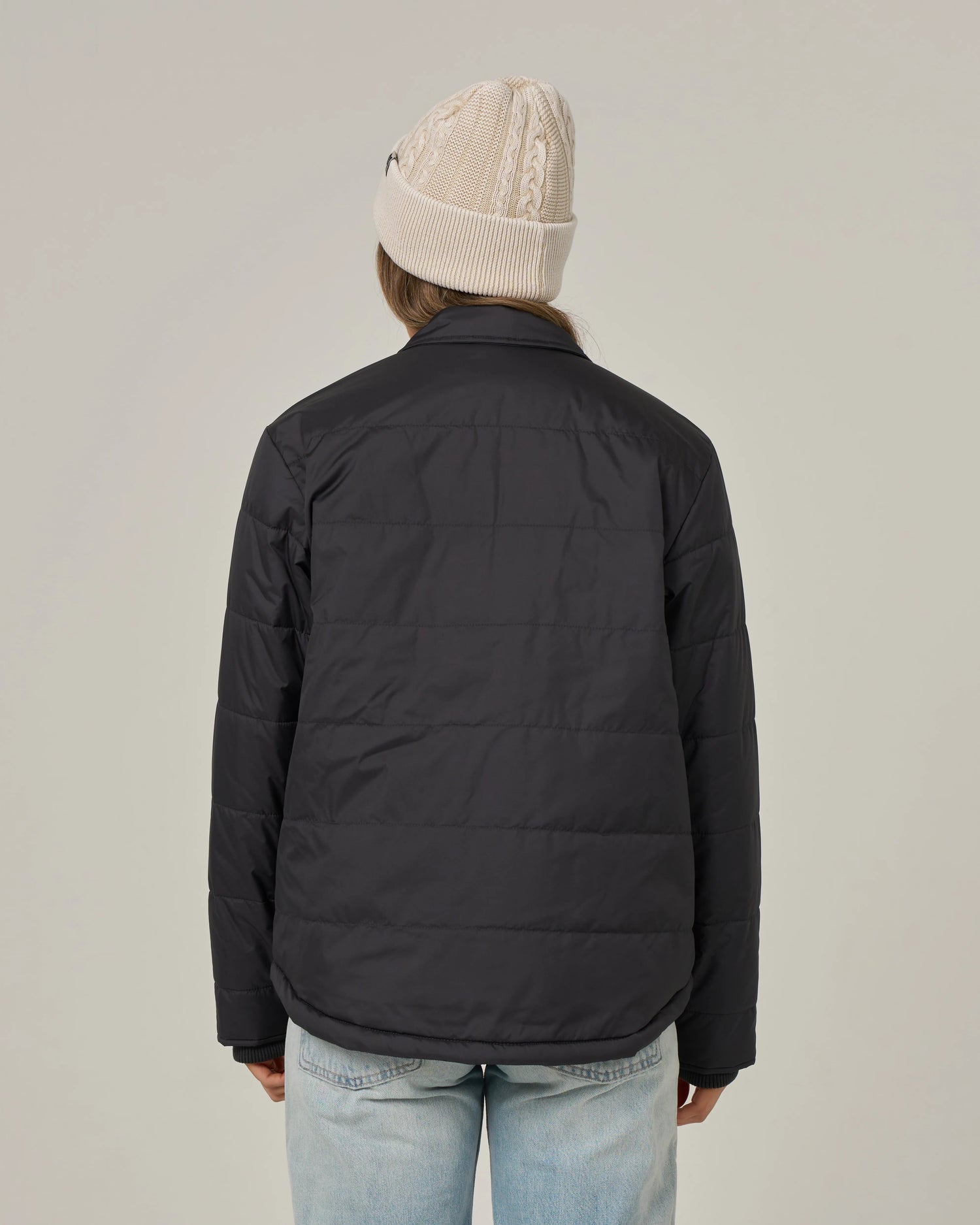 A person facing away, wearing a beige knit beanie, light blue jeans, and the Salty Crew Barrier Snap Jacket in black, stands against a plain light background.