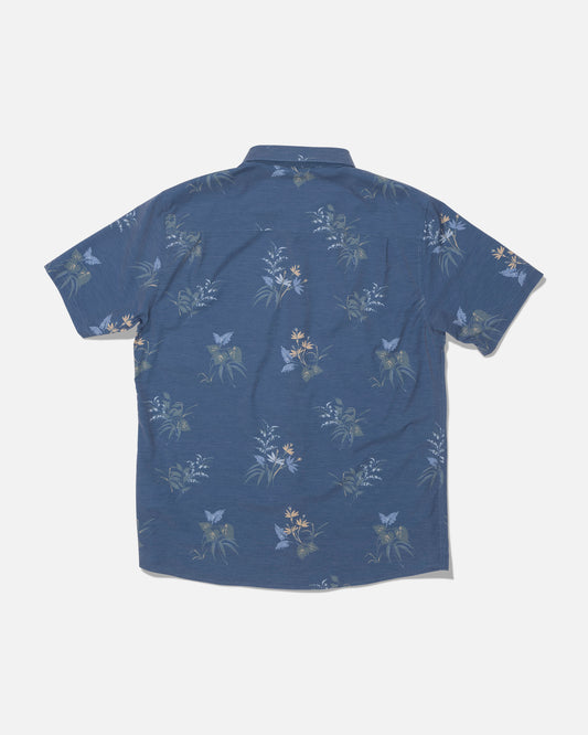 The Drifter UV Button Up - Navy by Salty Crew is a blue short-sleeve collared shirt, shown from the back against a white background. It features a subtle floral pattern in shades of light blue, green, and yellow. Made from moisture-wicking fabric with quick-dry properties, this shirt is designed to keep you cool and comfortable all day long.