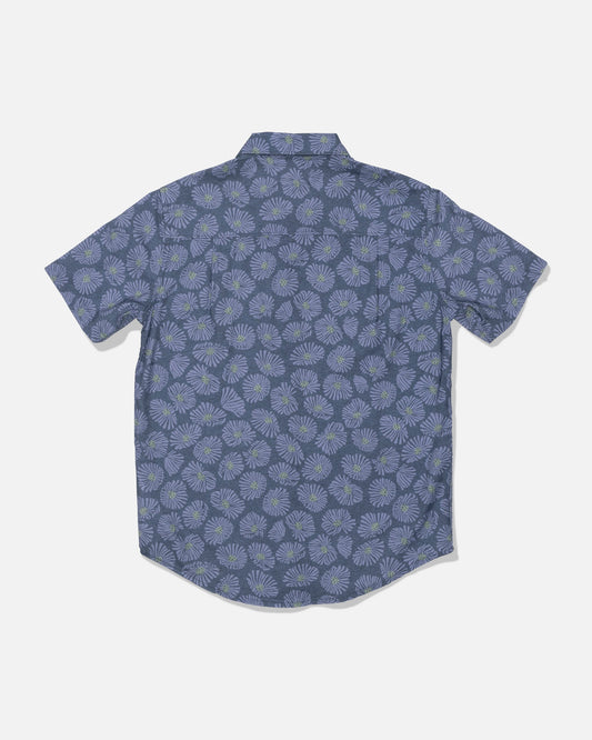 The Salty Crew Crew Boys Button Up in Hitch Blue is a short-sleeve, button-up shirt made from a soft cotton blend, featuring an allover print of light blue and green floral designs on a blue background. The shirt is displayed from the back against a plain white backdrop.