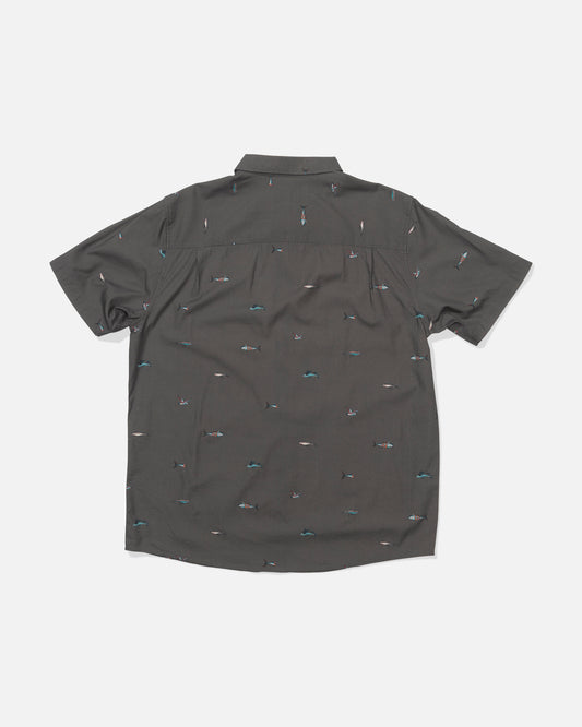 The Crew Button Up - Charcoal is shown laid flat, back facing up. This short-sleeve shirt features an allover print of small, colorful surfers and waves on a dark gray cotton blend fabric.