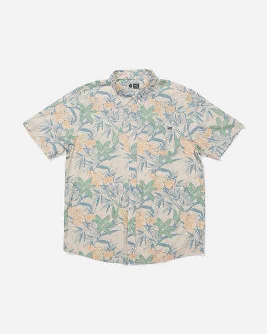 The Crew Button Up - Off White is a short-sleeve shirt featuring a light beige background and green, yellow, and blue floral leaf print. Made from a mid-weight cotton blend, it has a collar and a subtle woven chest label.
