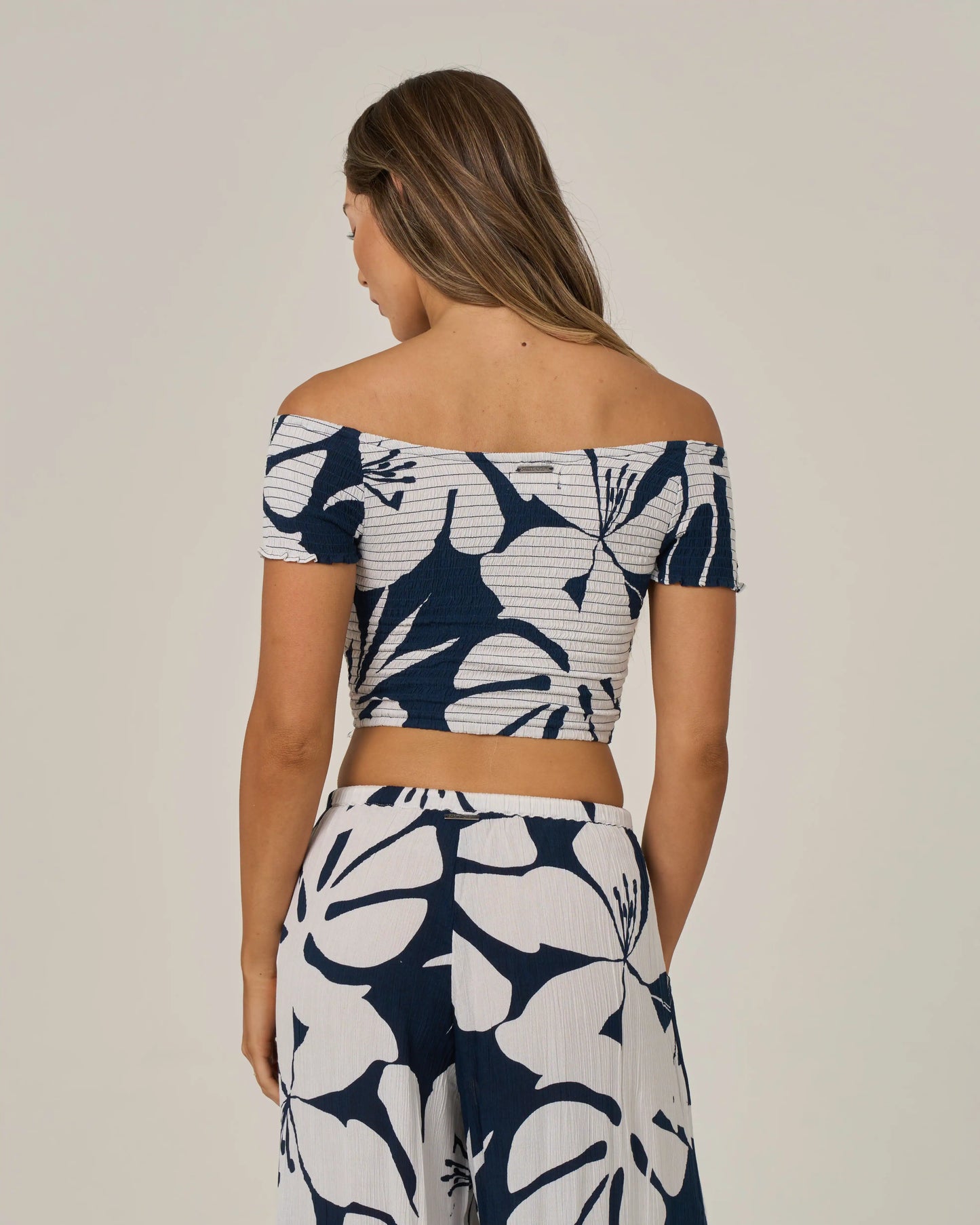 A woman faces away, wearing the Salty Crew Graphic Bloom Top in Dark Navy—an off-the-shoulder crop top with convertible sleeves—paired with matching wide-leg pants in a bold navy and white floral pattern, set against a plain background.