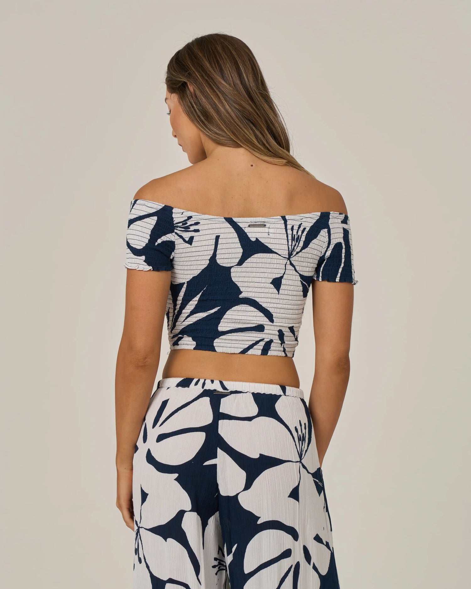 A woman faces away, wearing the Salty Crew Graphic Bloom Top in Dark Navy—an off-the-shoulder crop top with convertible sleeves—paired with matching wide-leg pants in a bold navy and white floral pattern, set against a plain background.