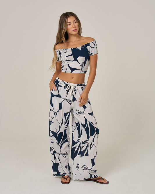 A woman stands barefoot on a plain background, wearing the Salty Crew Graphic Bloom Top in dark navy—a viscose, off-the-shoulder smocked top with convertible sleeves—paired with matching wide-leg pants featuring a navy and white floral print.