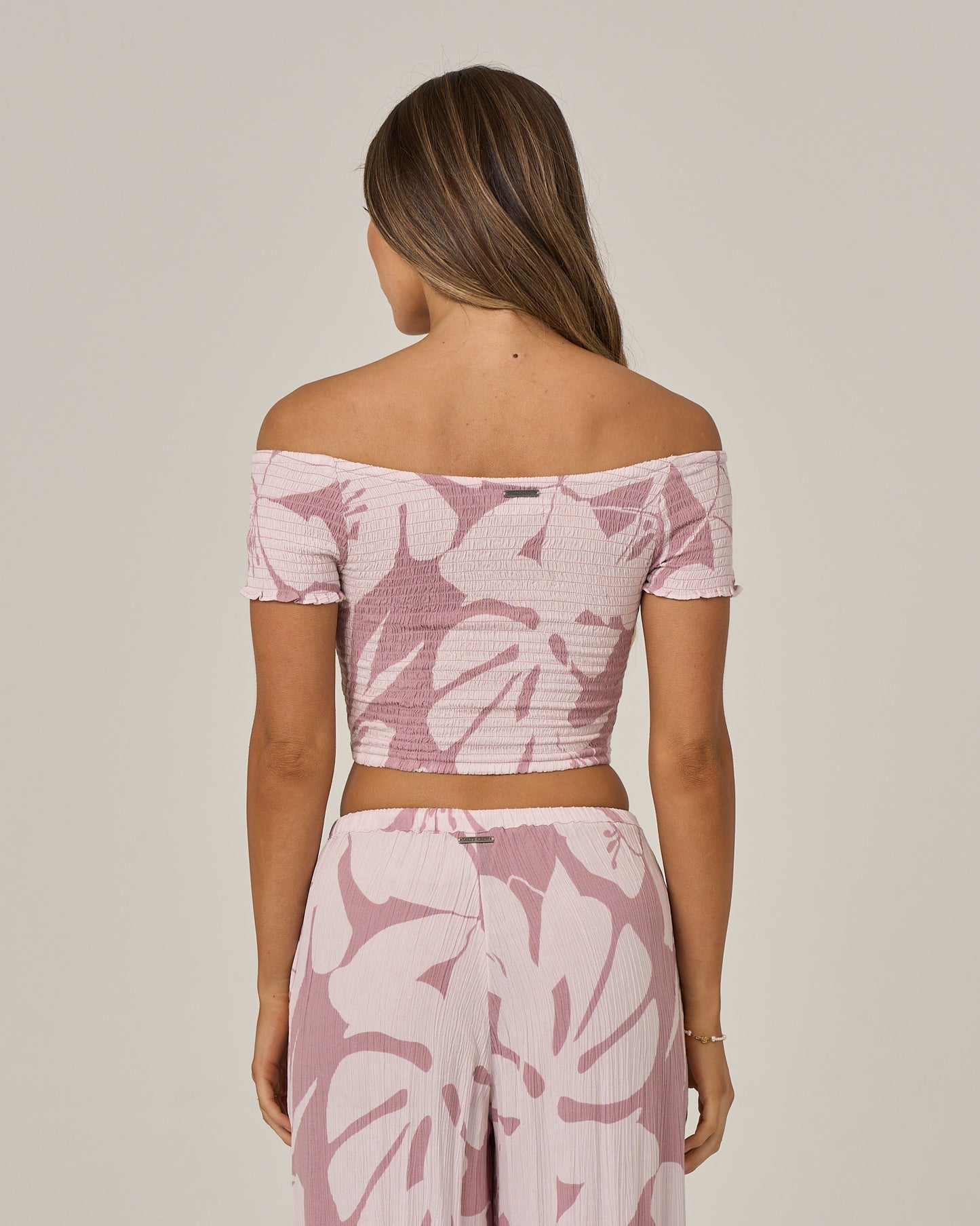 A woman stands facing away, wearing the Salty Crew Graphic Bloom Top in Faded Mauve—a pink and white off-the-shoulder style with a bold leaf print. The viscose smocked top features convertible sleeves for versatile styling, paired with matching flowy pants. Her long hair is down and her arms rest relaxed at her sides against a plain background.