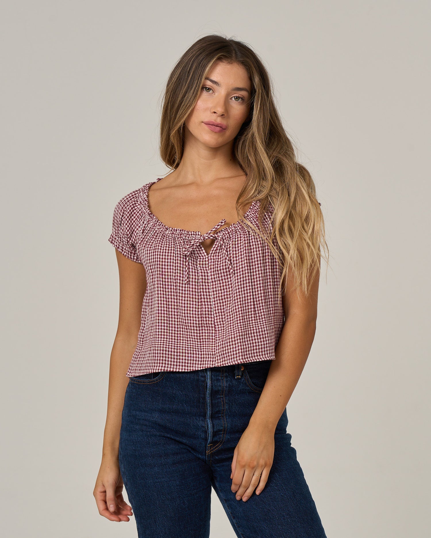 A woman with long, wavy hair wears the “Fresh Catch - Maroon” cotton double gauze top with ruffle sleeves by Salty Crew and dark blue jeans, standing against a plain light background.