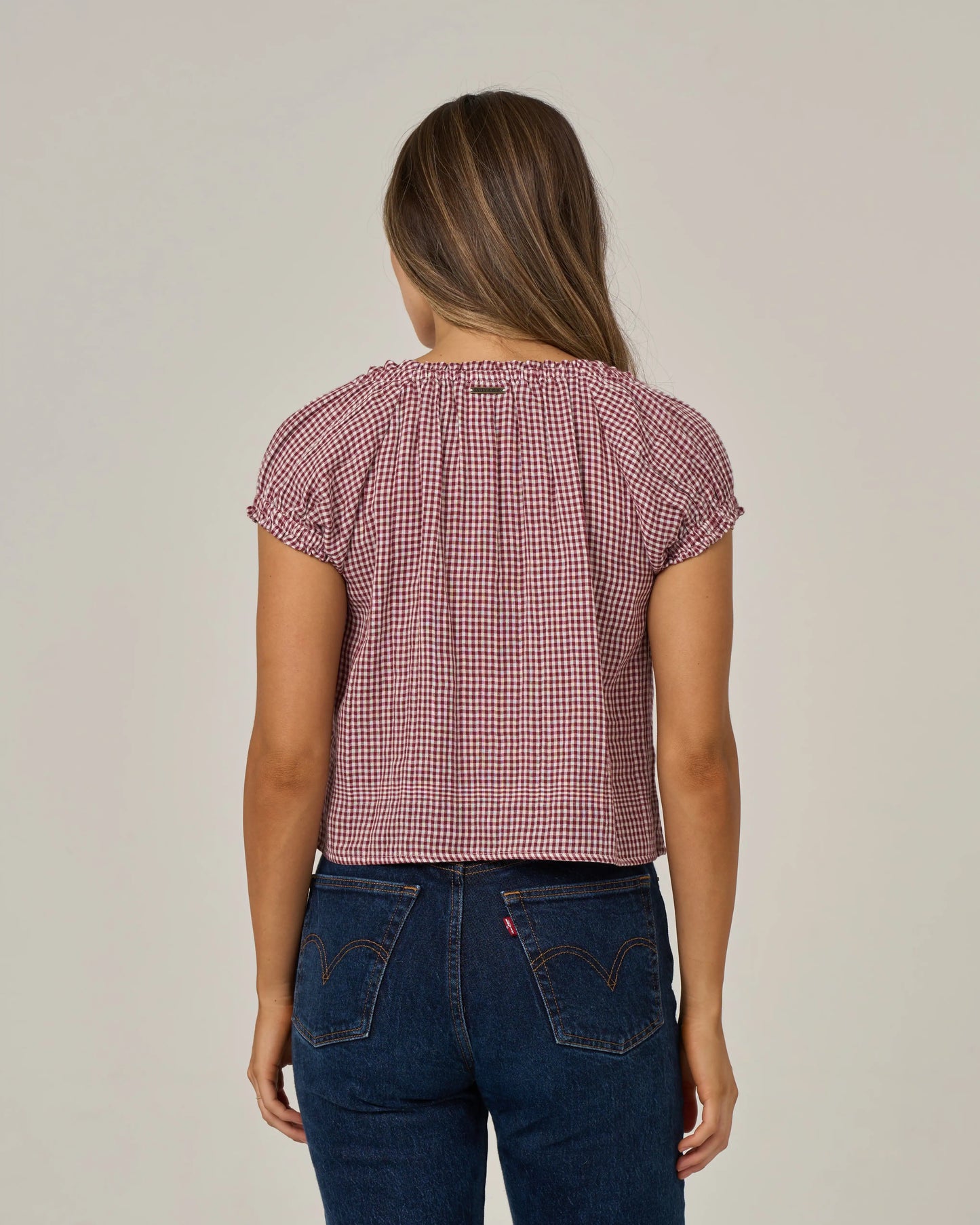 A woman stands facing away, wearing the Salty Crew Fresh Catch - Maroon cotton double gauze top featuring short sleeves and a red and white checkered pattern, paired with dark blue jeans accented by visible stitching and back pockets. Her straight brown hair is worn down, and the background is plain and neutral.