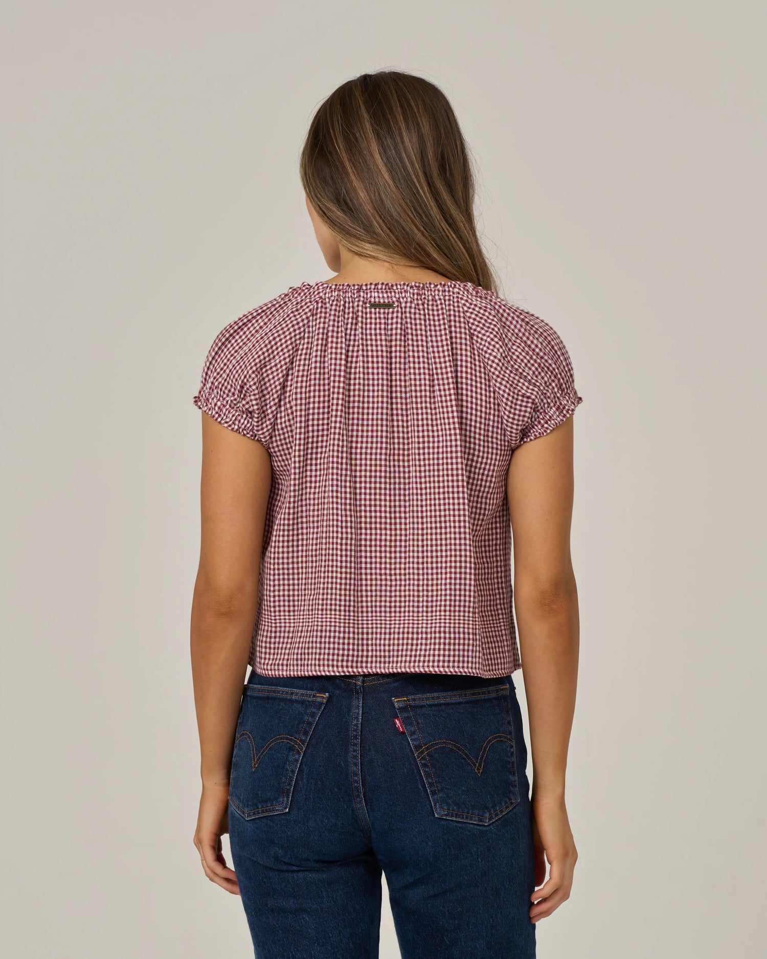 A woman stands facing away, wearing the Salty Crew Fresh Catch - Maroon cotton double gauze top featuring short sleeves and a red and white checkered pattern, paired with dark blue jeans accented by visible stitching and back pockets. Her straight brown hair is worn down, and the background is plain and neutral.