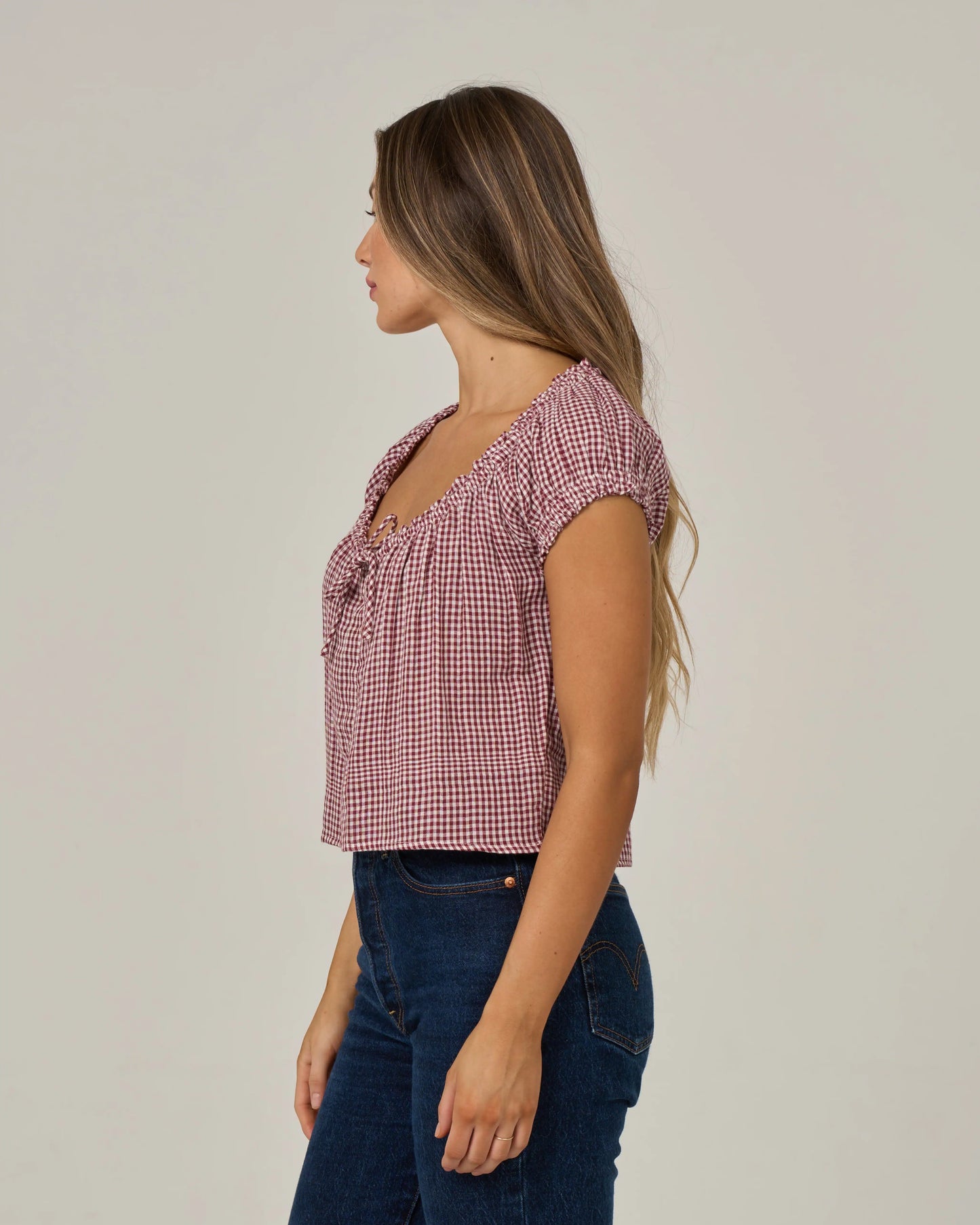 A woman with long hair stands in profile against a plain background, wearing the Salty Crew Fresh Catch - Maroon top—a ruffle sleeve, red and white checkered cotton double gauze shirt—paired with blue jeans.