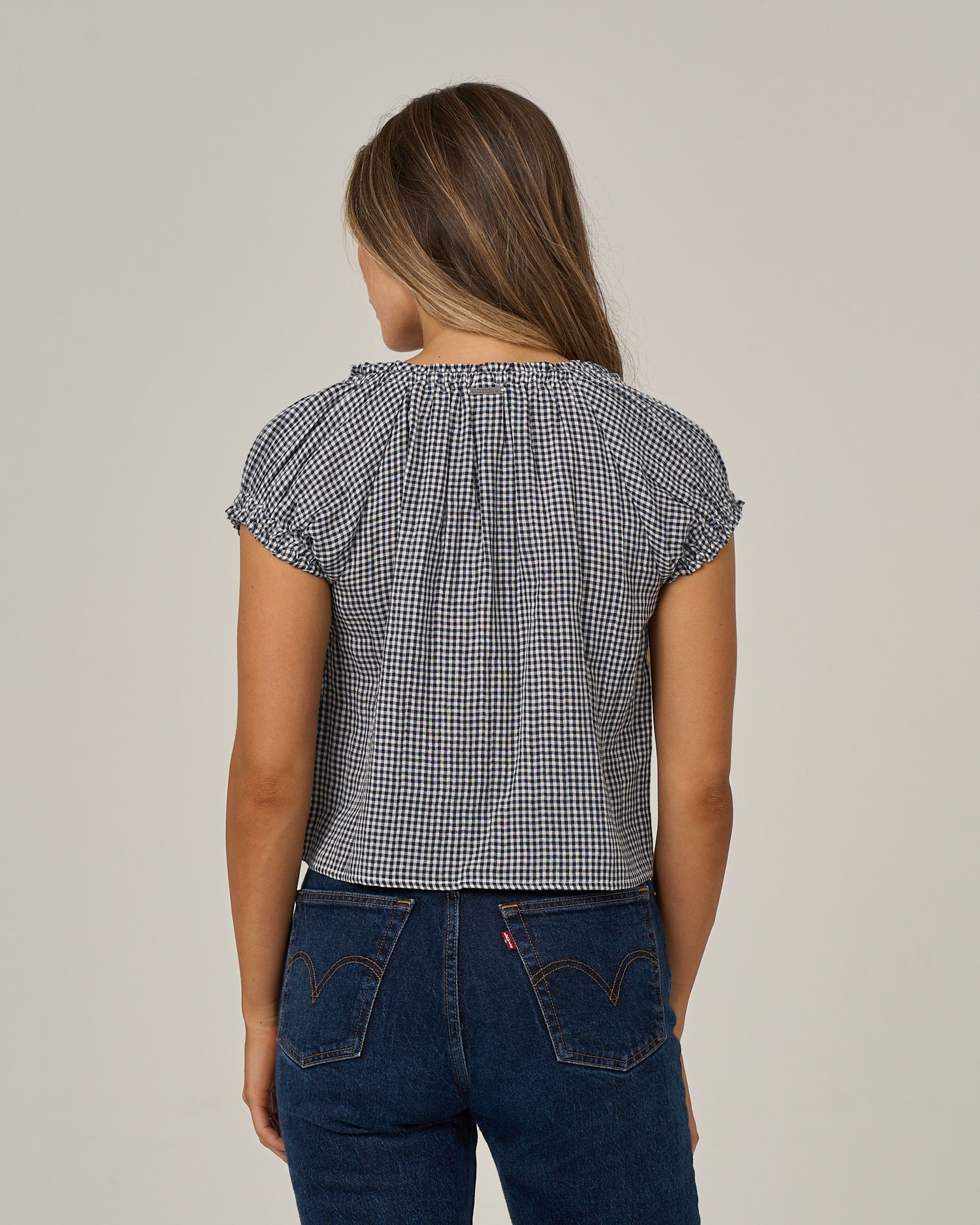 A person with long brown hair is standing facing away from the camera against a plain background, wearing Salty Crew’s “Fresh Catch” blouse in True Navy and blue jeans.