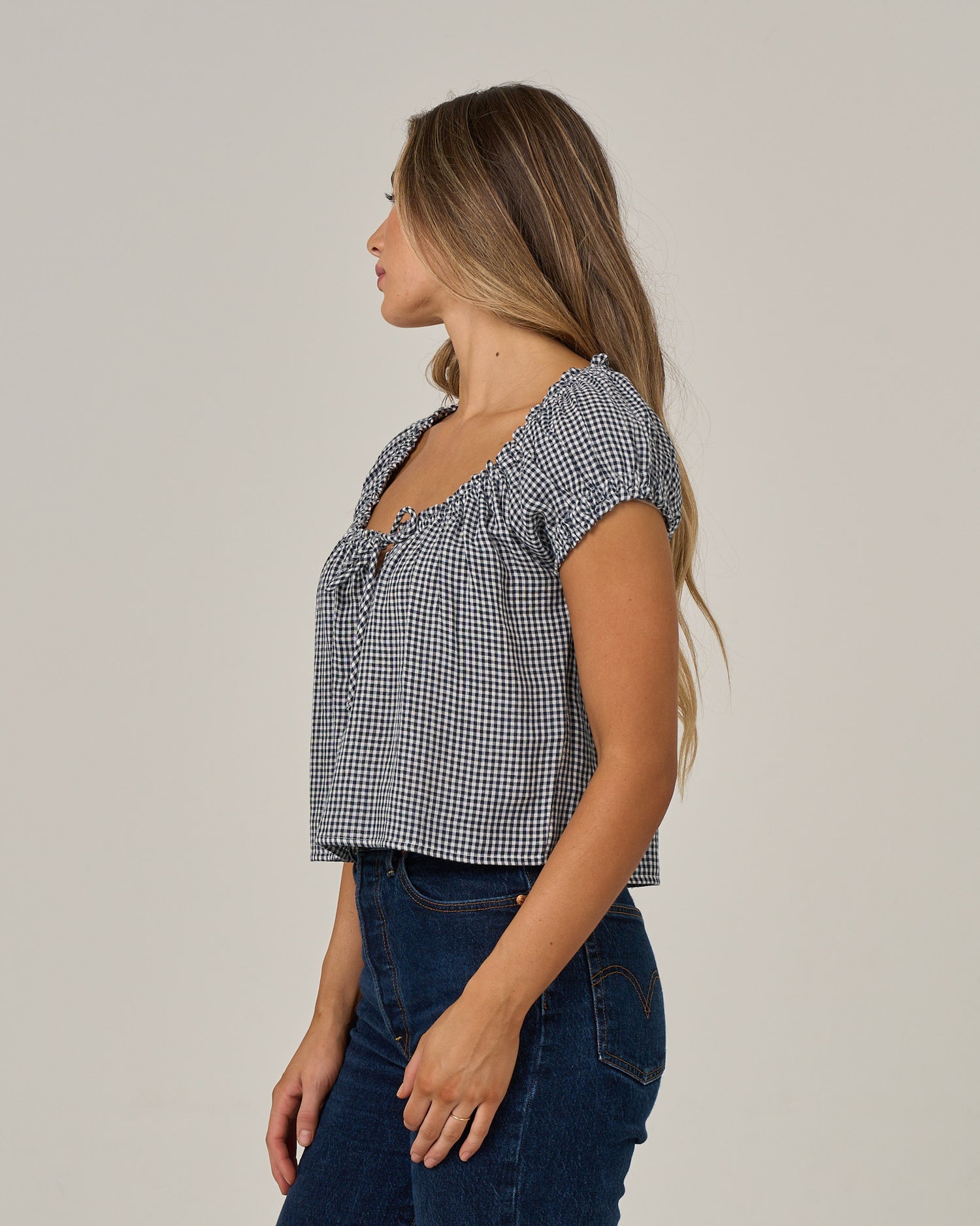 A woman with long hair, wearing the Salty Crew “Fresh Catch - True Navy” blue and white checkered cotton double gauze top with ruffle sleeves and dark blue jeans, stands sideways against a plain light background.