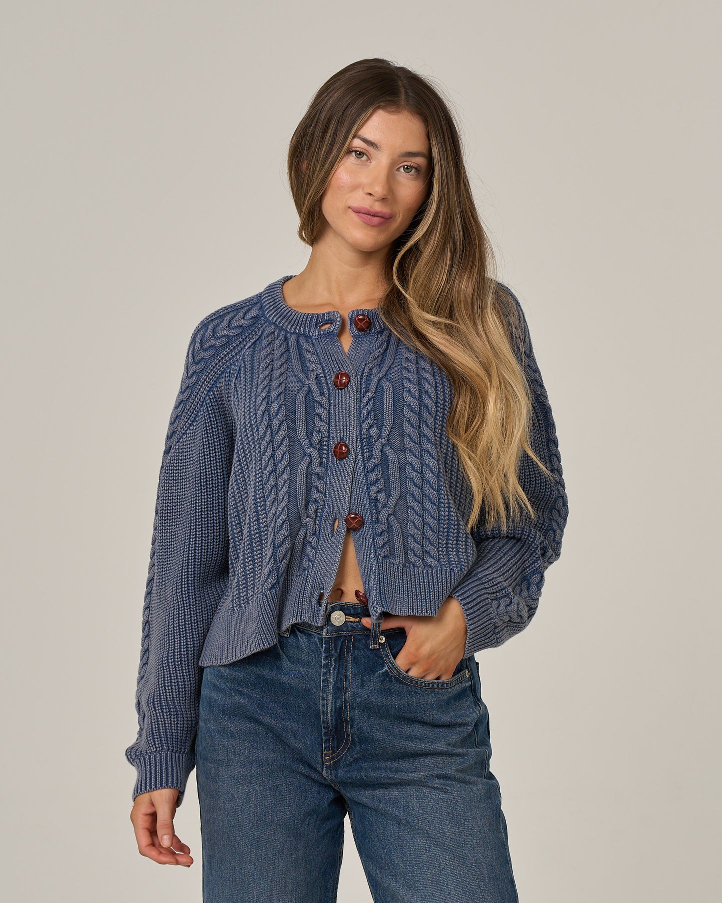 A woman with long, wavy hair wears the Salty Crew Fisherman Cardigan in Flint—crafted from soft cotton sweater material with brown buttons—paired with blue jeans, standing against a plain light background.